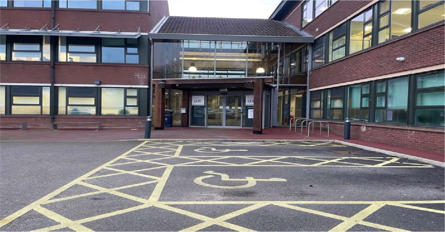 Exterior view of a building with a glass entrance, brick walls, and a marked accessible parking space.