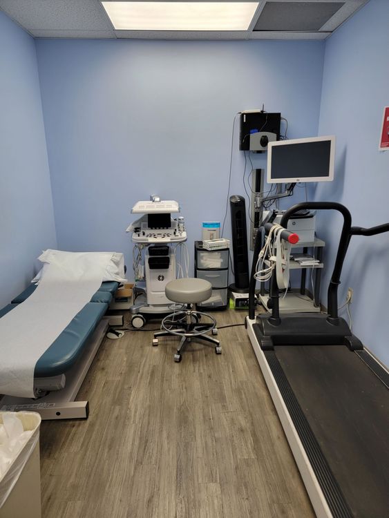 Small medical exam room with treatment chair, equipment cart, monitor, and treadmill against blue walls.