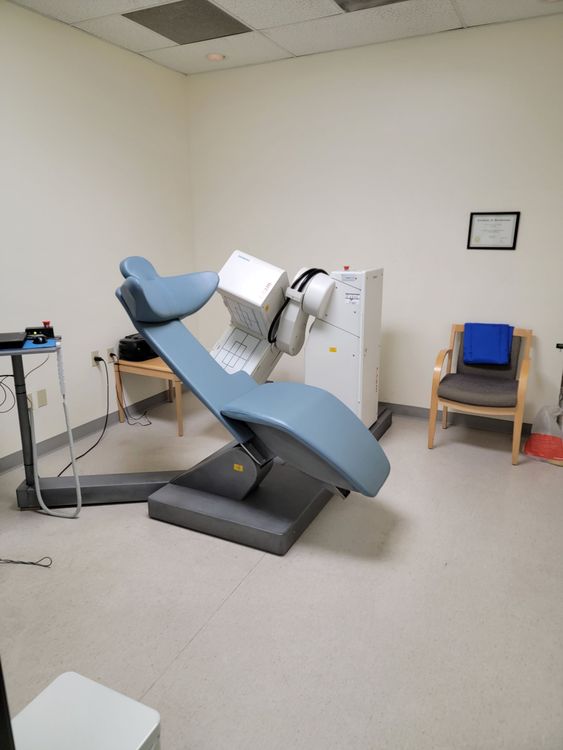 Tilted blue medical chair in a clinical room, with equipment beside it and chairs against the wall.