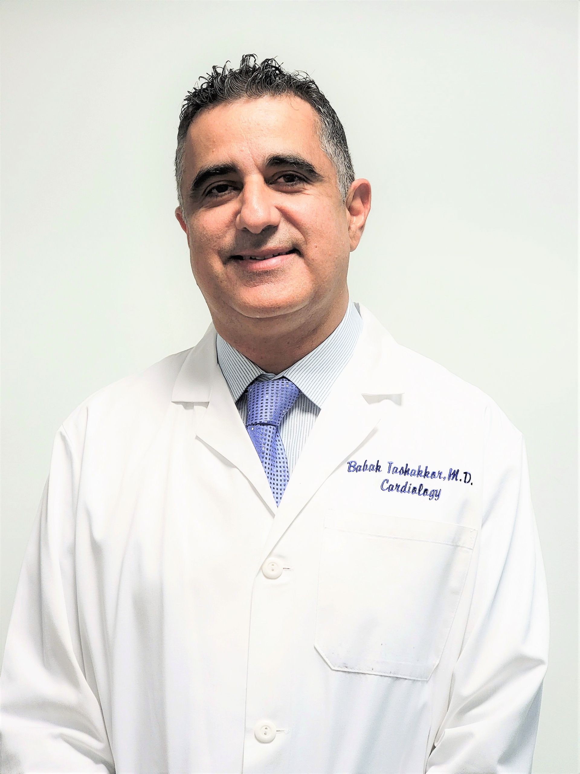 Doctor in a white coat and patterned tie, standing against a plain light background and smiling.