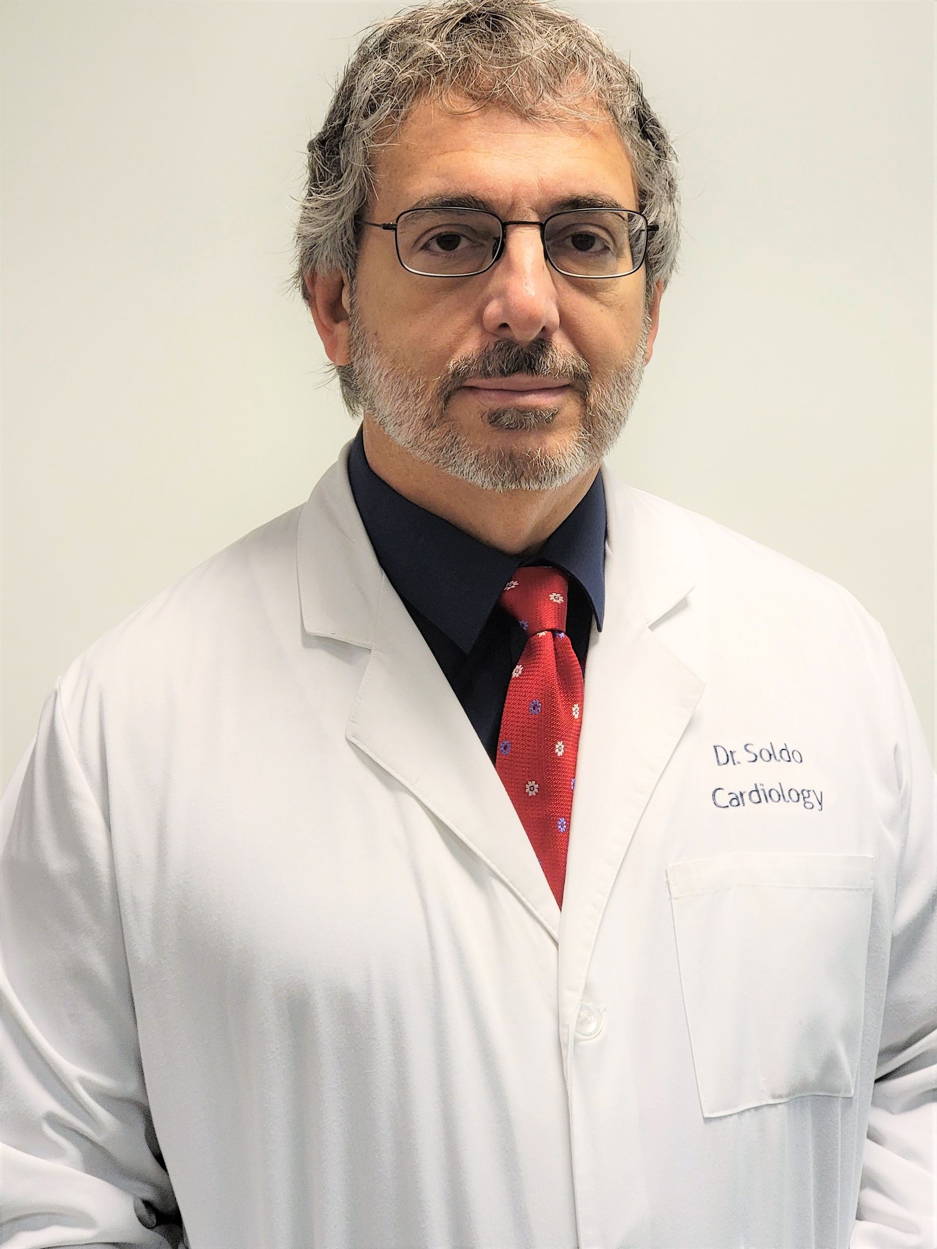 Doctor in a white lab coat, glasses, and red tie, standing against a plain light wall.