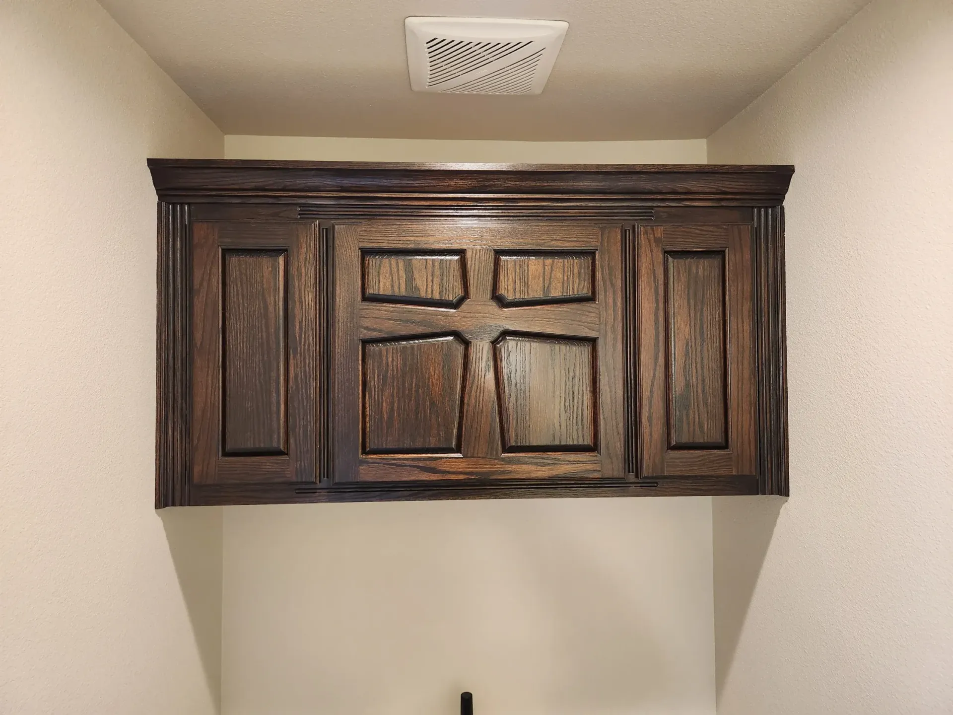 Dark wood cabinet mounted above a cream-colored wall in a room. A ventilation fan is above the cabinet.