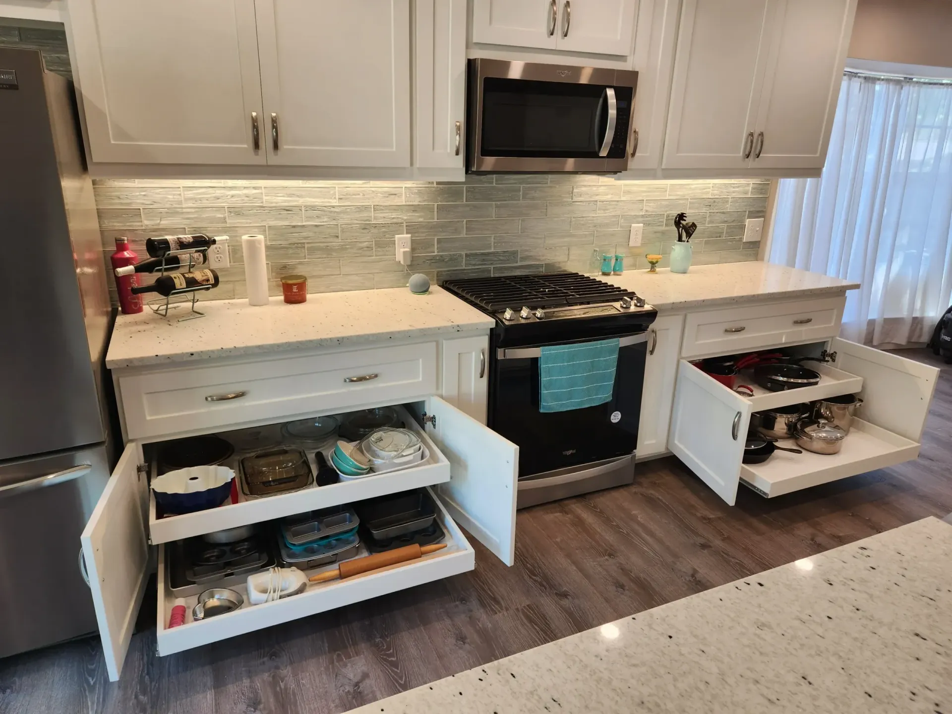 White kitchen cabinets with pull-out drawers, stove, microwave, and open storage. Light countertops, wood floor.