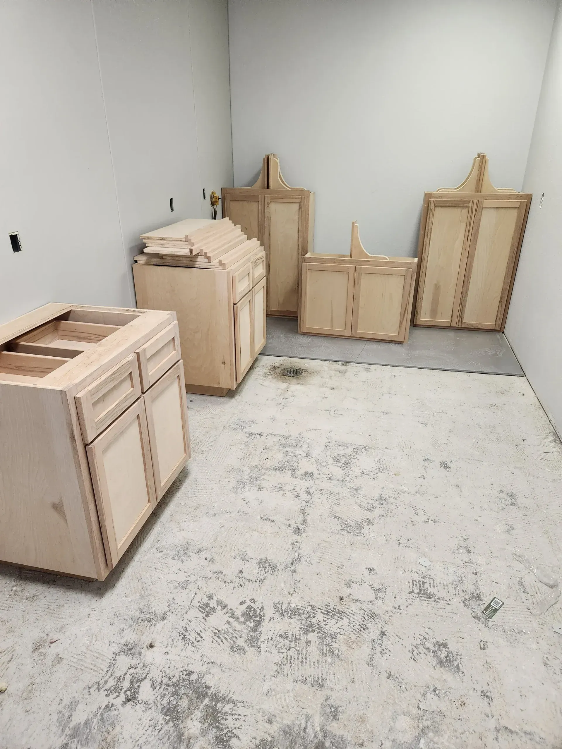 Unfinished wood kitchen cabinets in a room with concrete floor and white walls, awaiting installation.