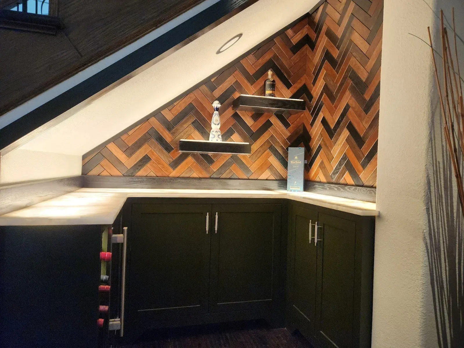 Dark green cabinets beneath angled wood-patterned wall, white counter, shelves with bottles.