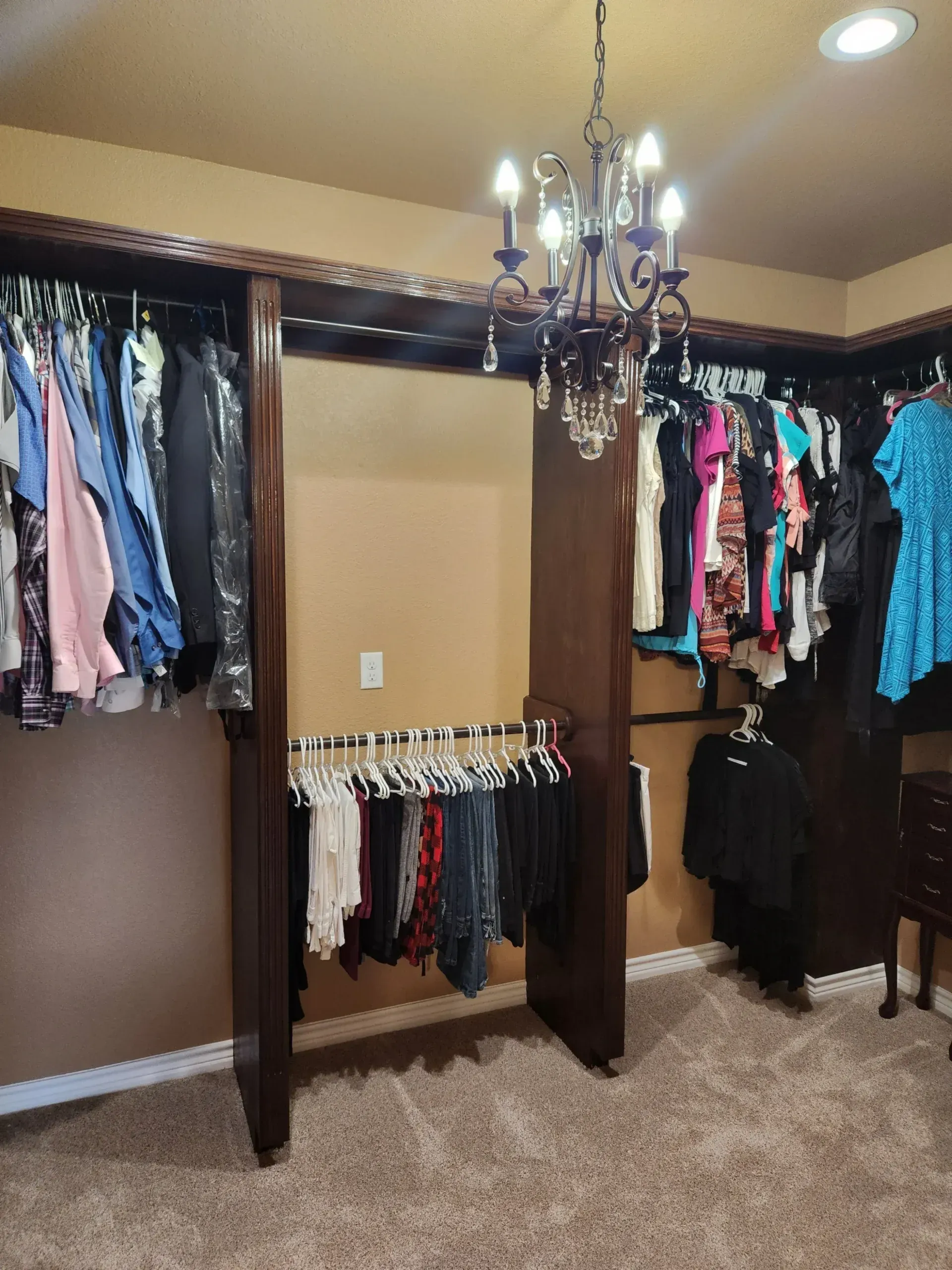 Walk-in closet with dark wood trim, filled with hanging clothes. A chandelier hangs from the ceiling.