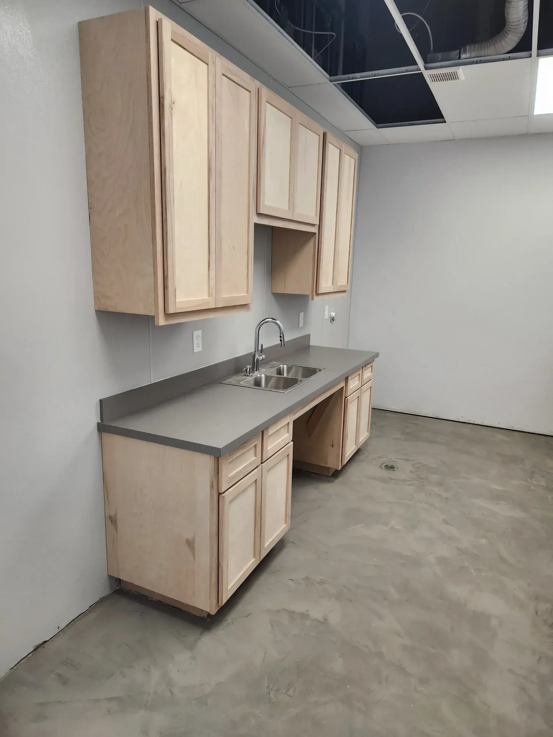 Kitchenette with light wood cabinets, gray countertop, and stainless steel sink.