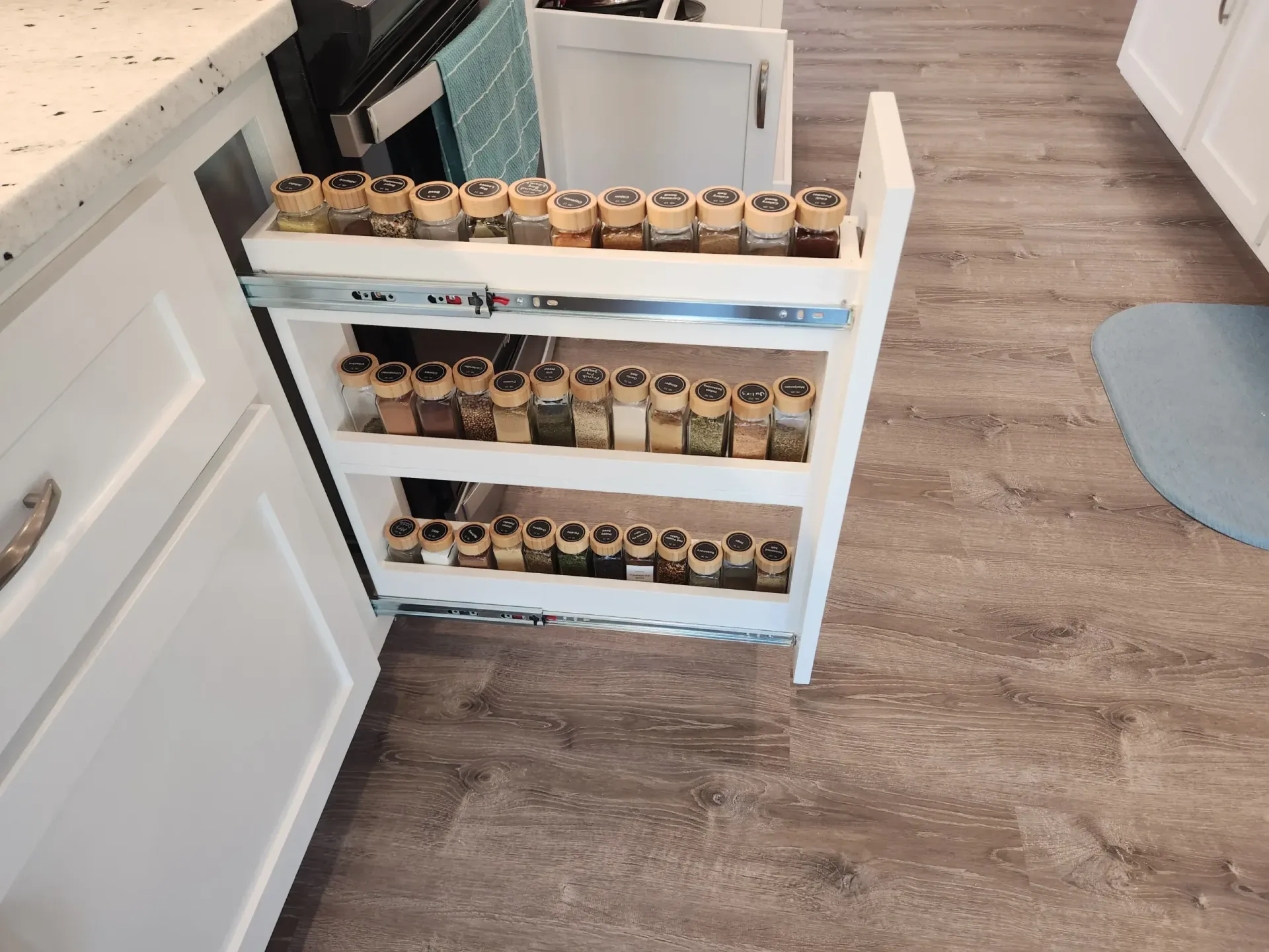 White pull-out spice rack with various spice jars inside a white cabinet, next to a countertop.