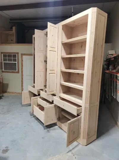 Wooden unfinished custom bookshelf with open doors and drawers in a workshop.