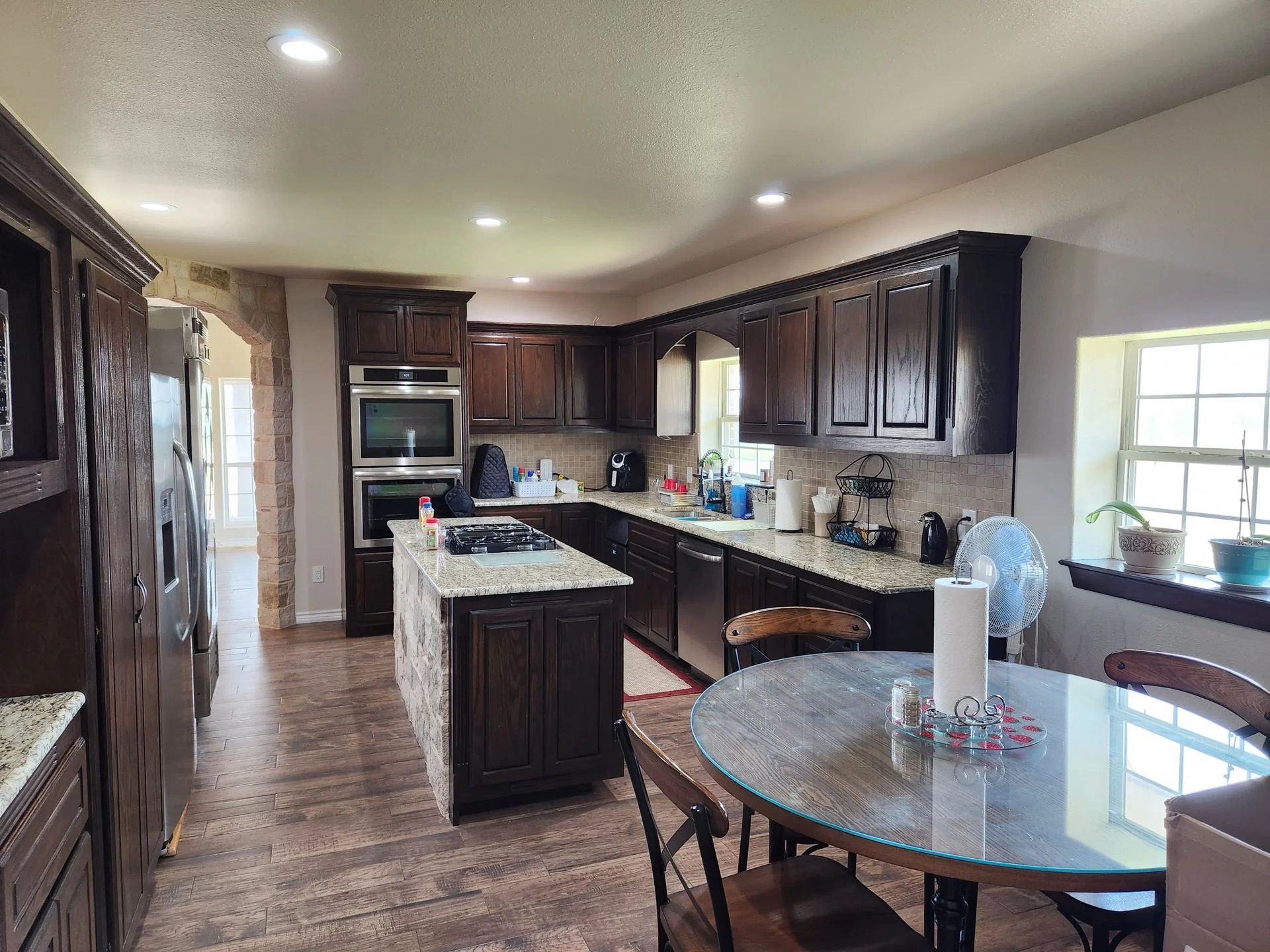 Dark-brown kitchen with island, table, and appliances. Wooden floor. Bright lighting.