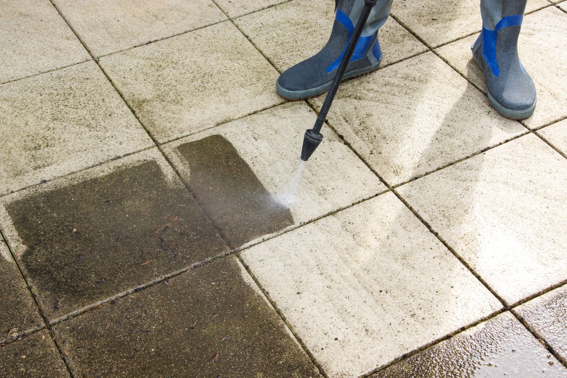 Person pressure-washing dirty tiled pavement with a spray nozzle, cleaning a dark stain.