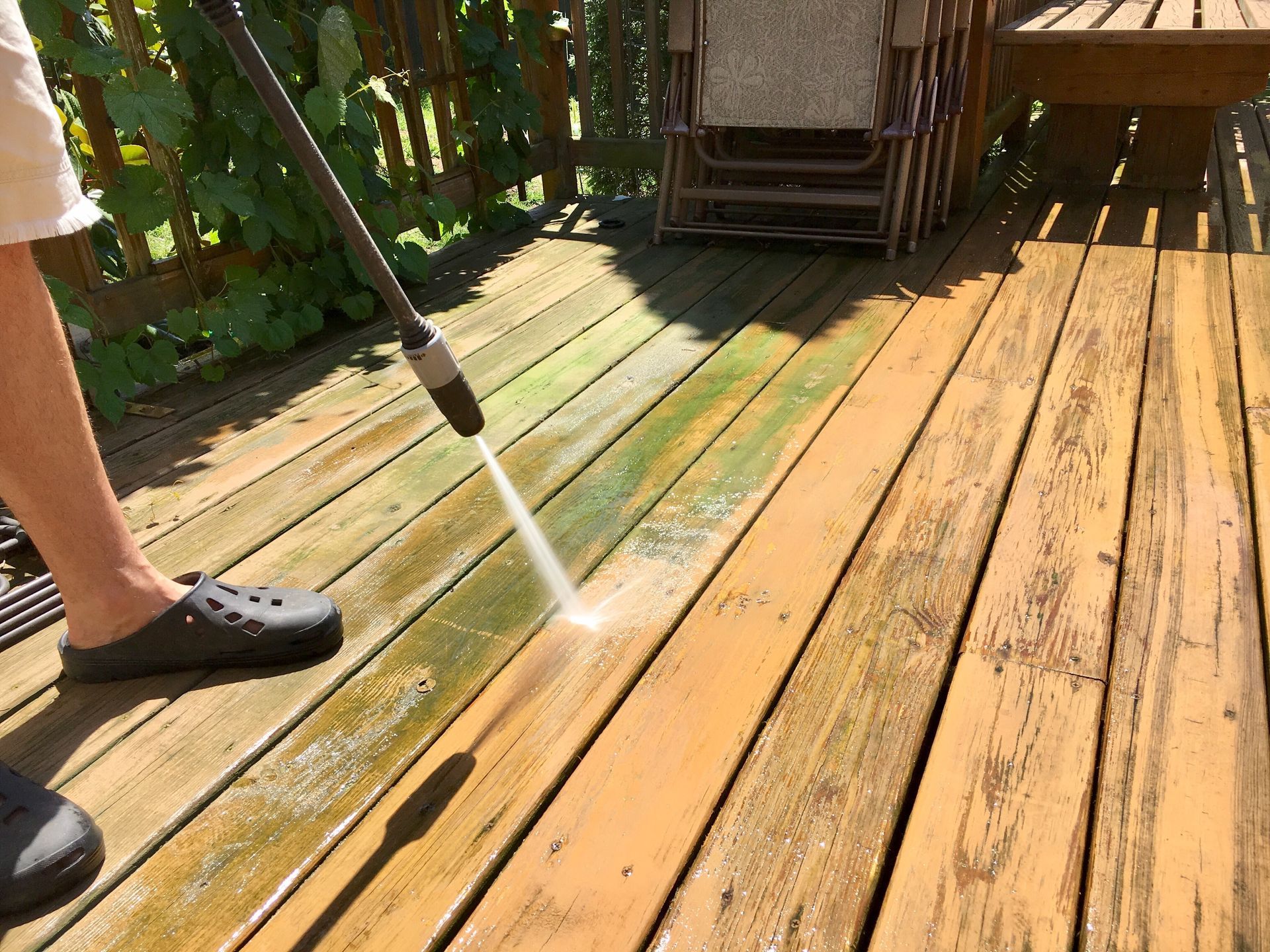 Person pressure-washing a sunlit wooden deck with green algae stains