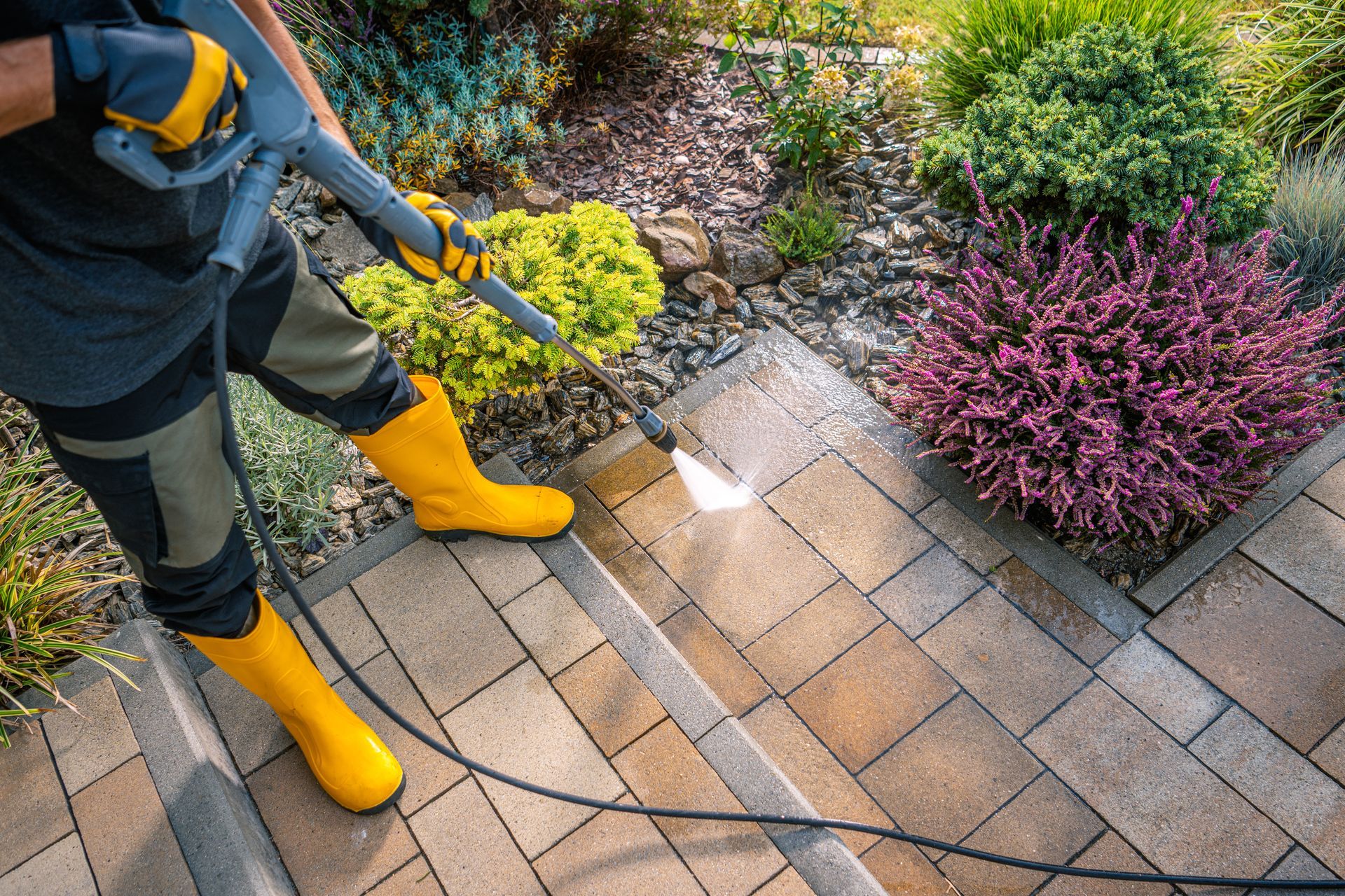 Person using a pressure washer on a stone patio beside colorful garden shrubs