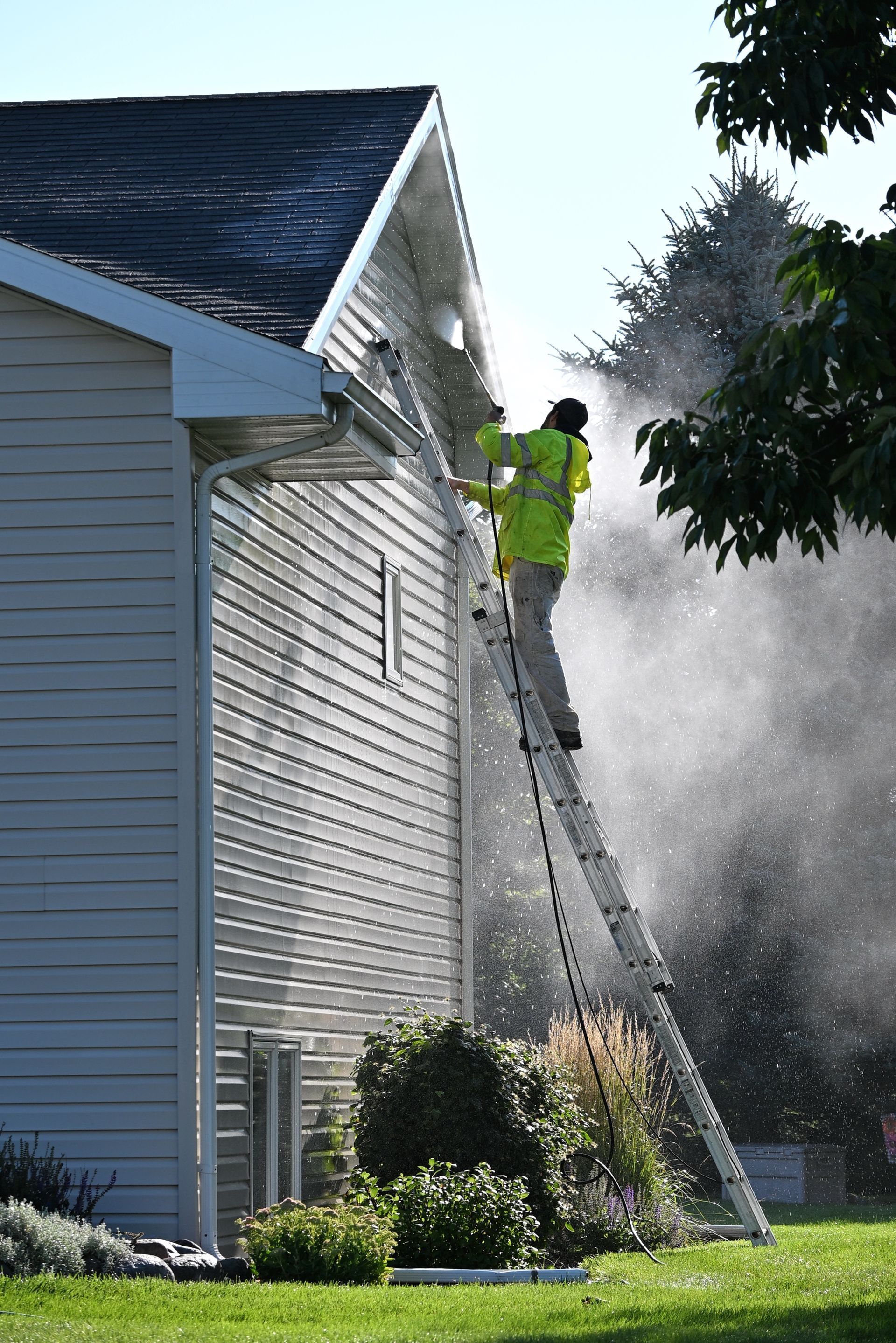 Worker pressure-washing the side of a house from a ladder, with water spray and green lawn below