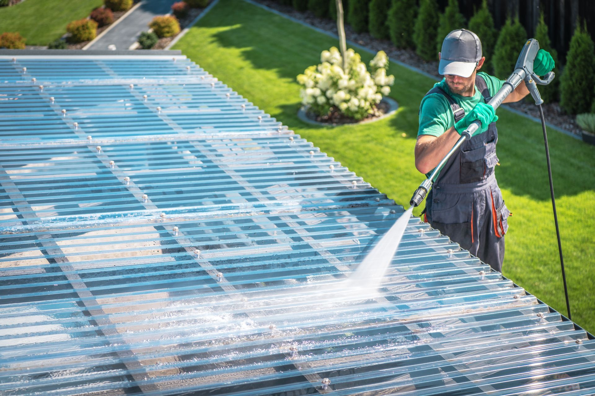 Worker pressure-washing a large roof beside a green lawn, wearing safety gear and holding a hose