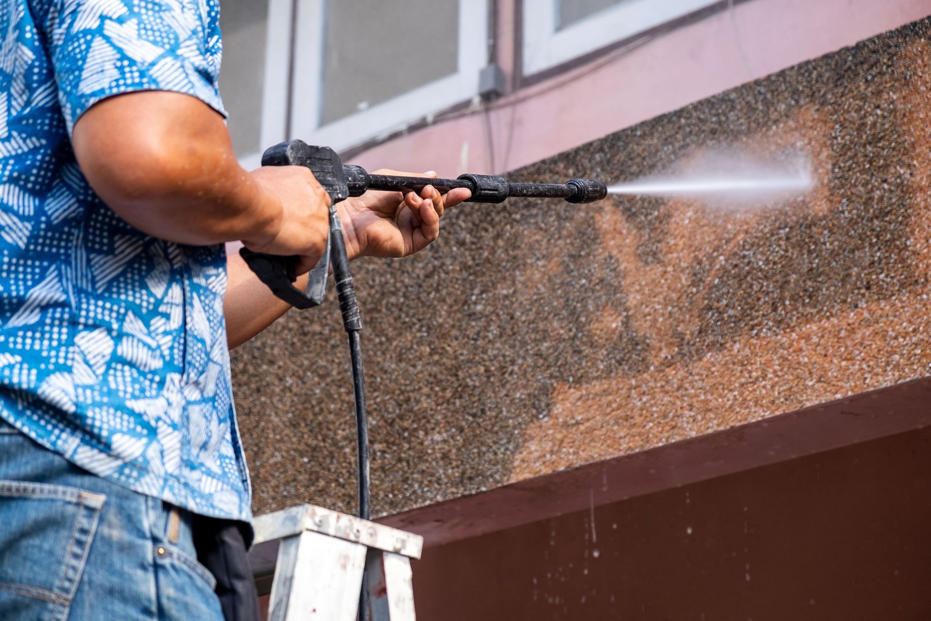 Worker pressure-washing a brown exterior wall on a ladder