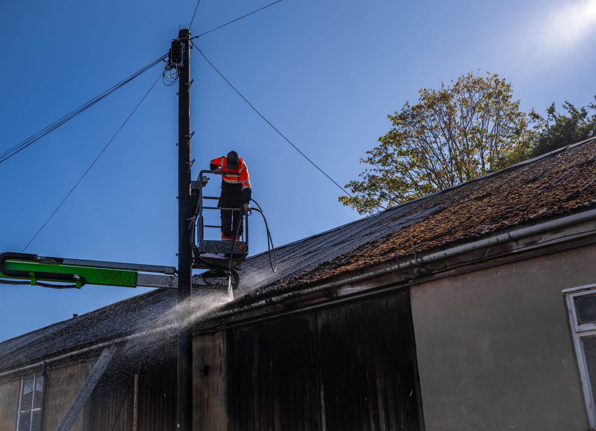 Worker pressure-washing a roof from a lift beside a utility pole under a blue sky
