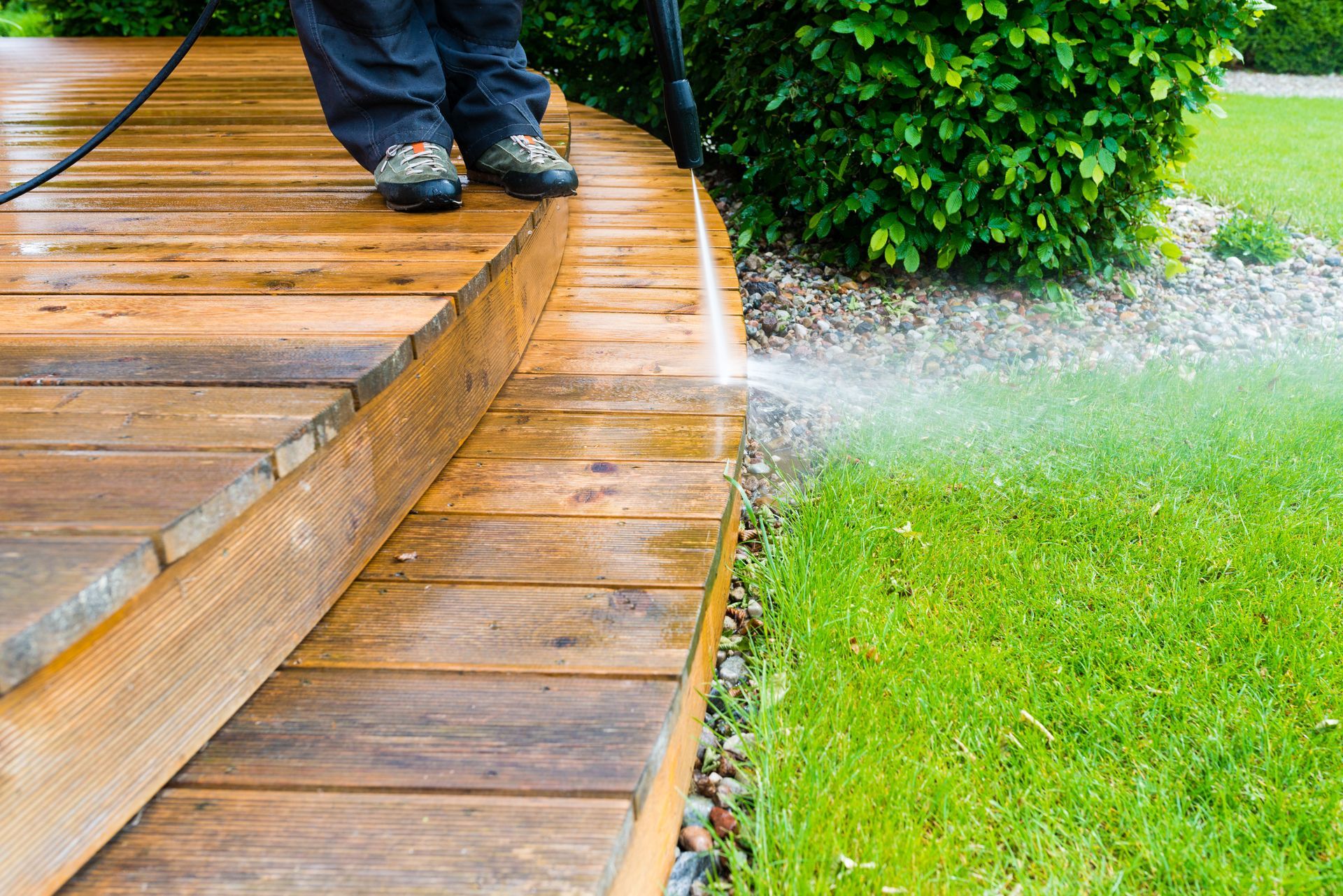 Person walking on a wet wooden deck beside a green lawn and bush after rain