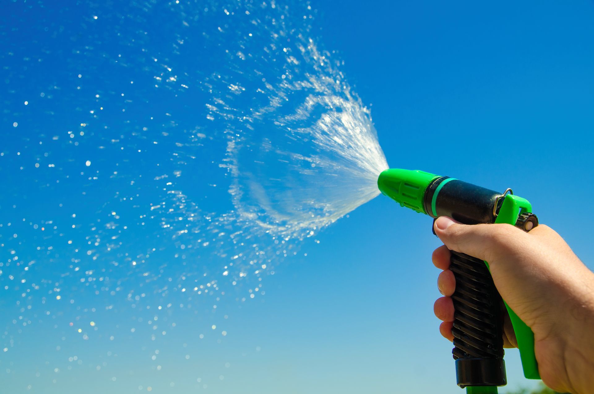 Hand holding a green hose nozzle spraying water into a clear blue sky