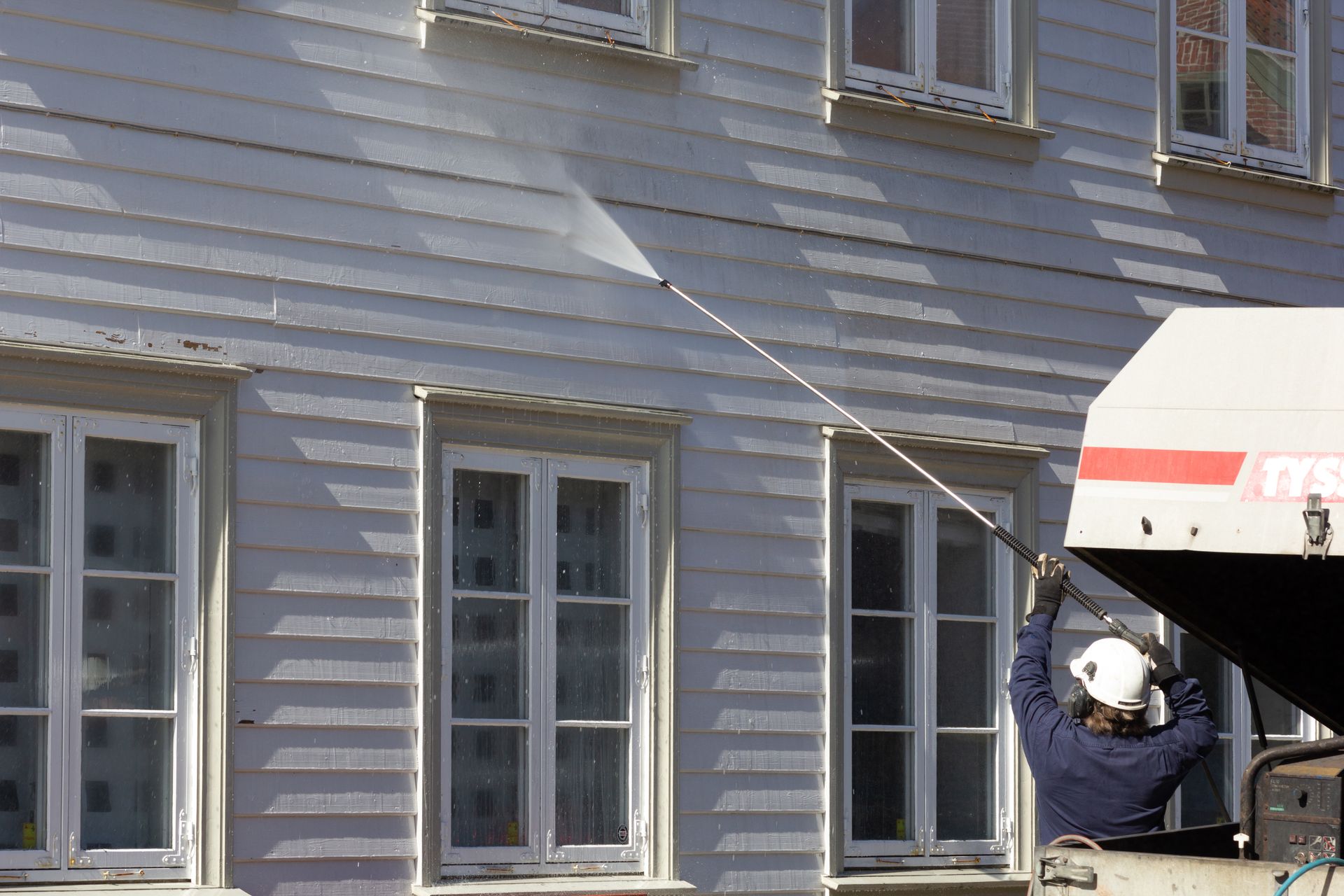 Worker cleaning house siding with a long pole next to a window and utility trailer