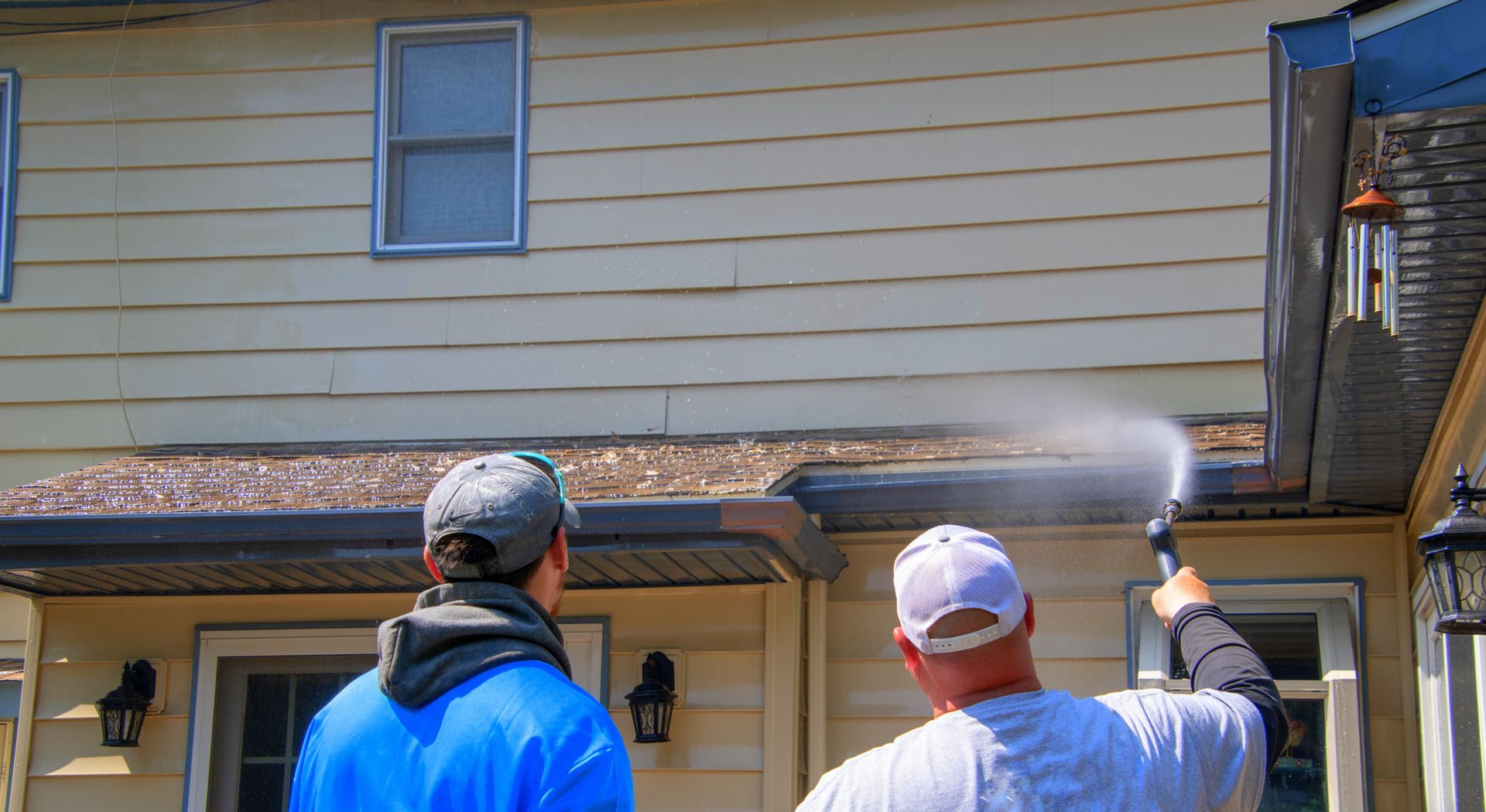 Two workers spray water on a tan house’s upper siding and eaves from below.