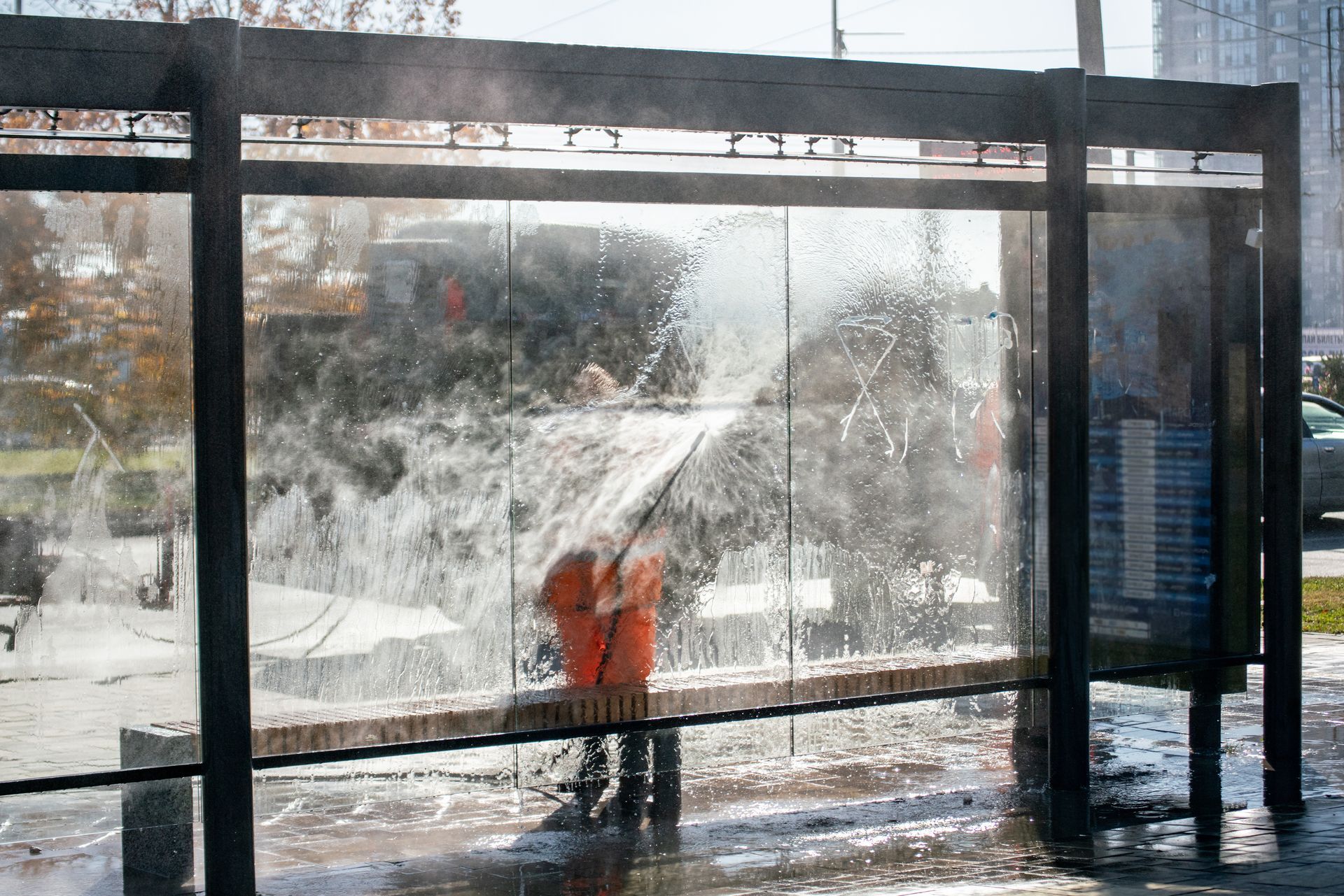 Person in orange using a snow blower outside a glass bus shelter in a snowy scene