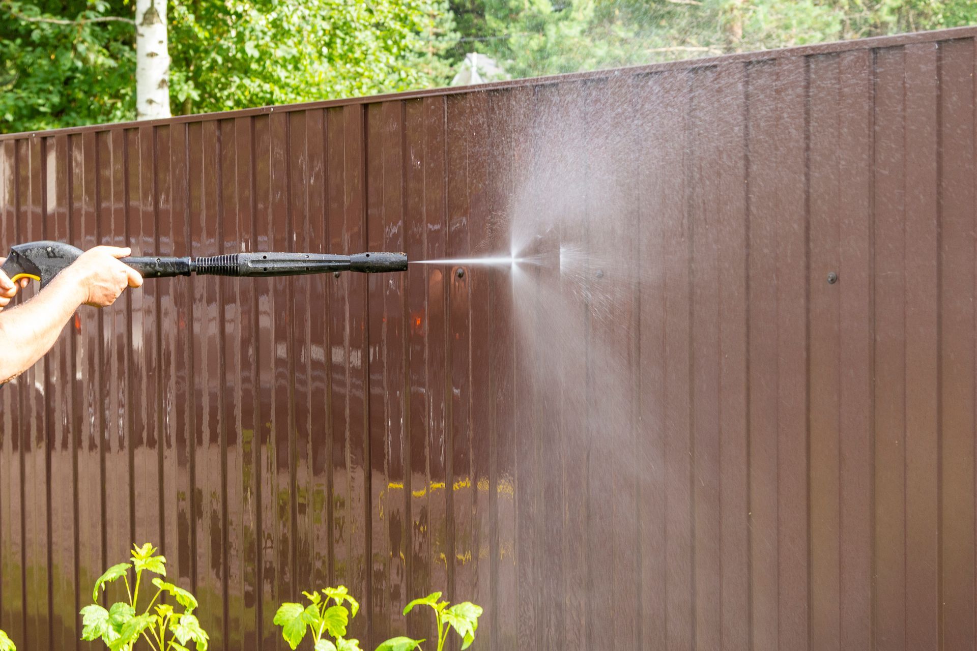 Person pressure-washing a brown wooden fence in a yard