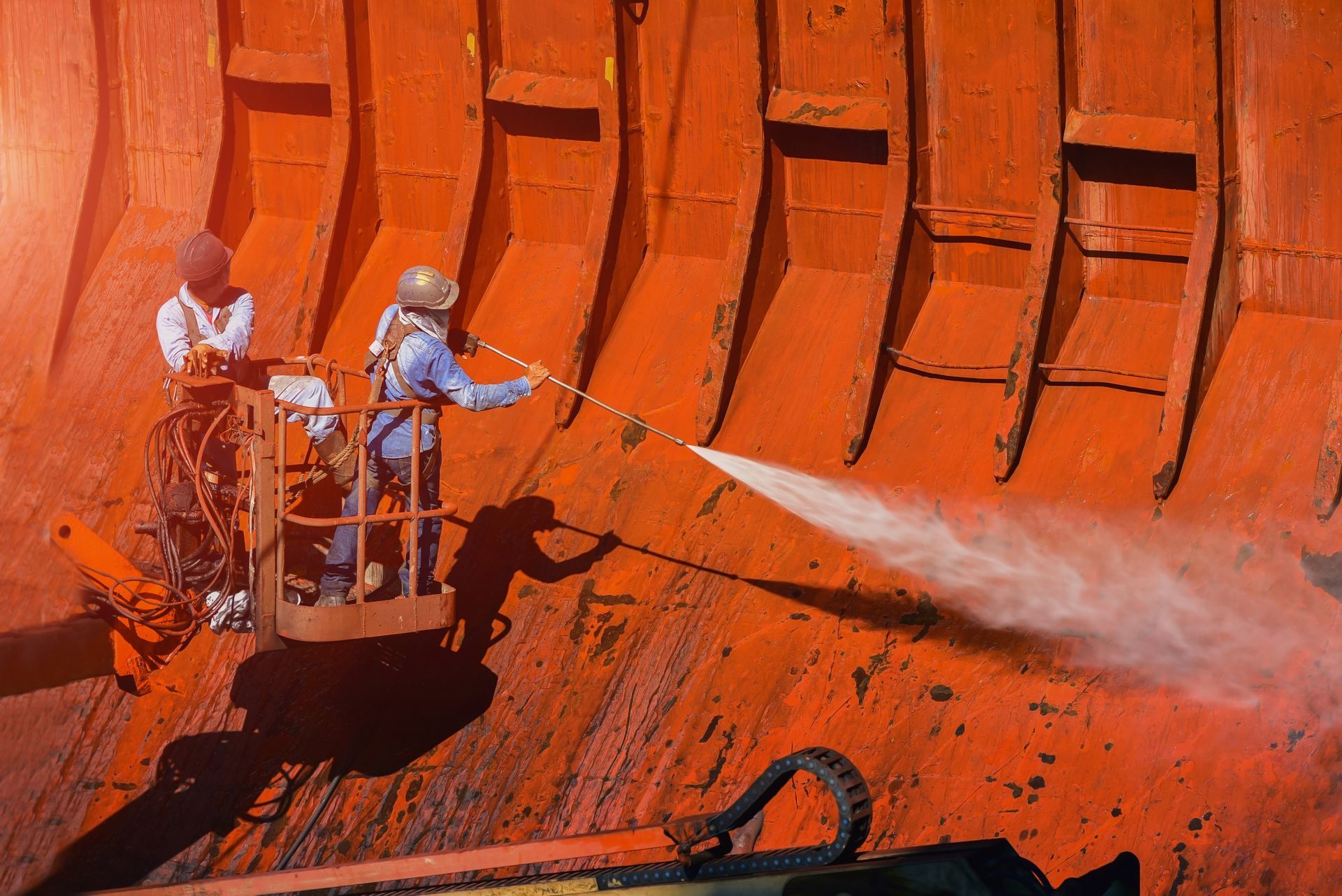 Workers spraying orange industrial tank interior with a high-pressure hose from a lift platform