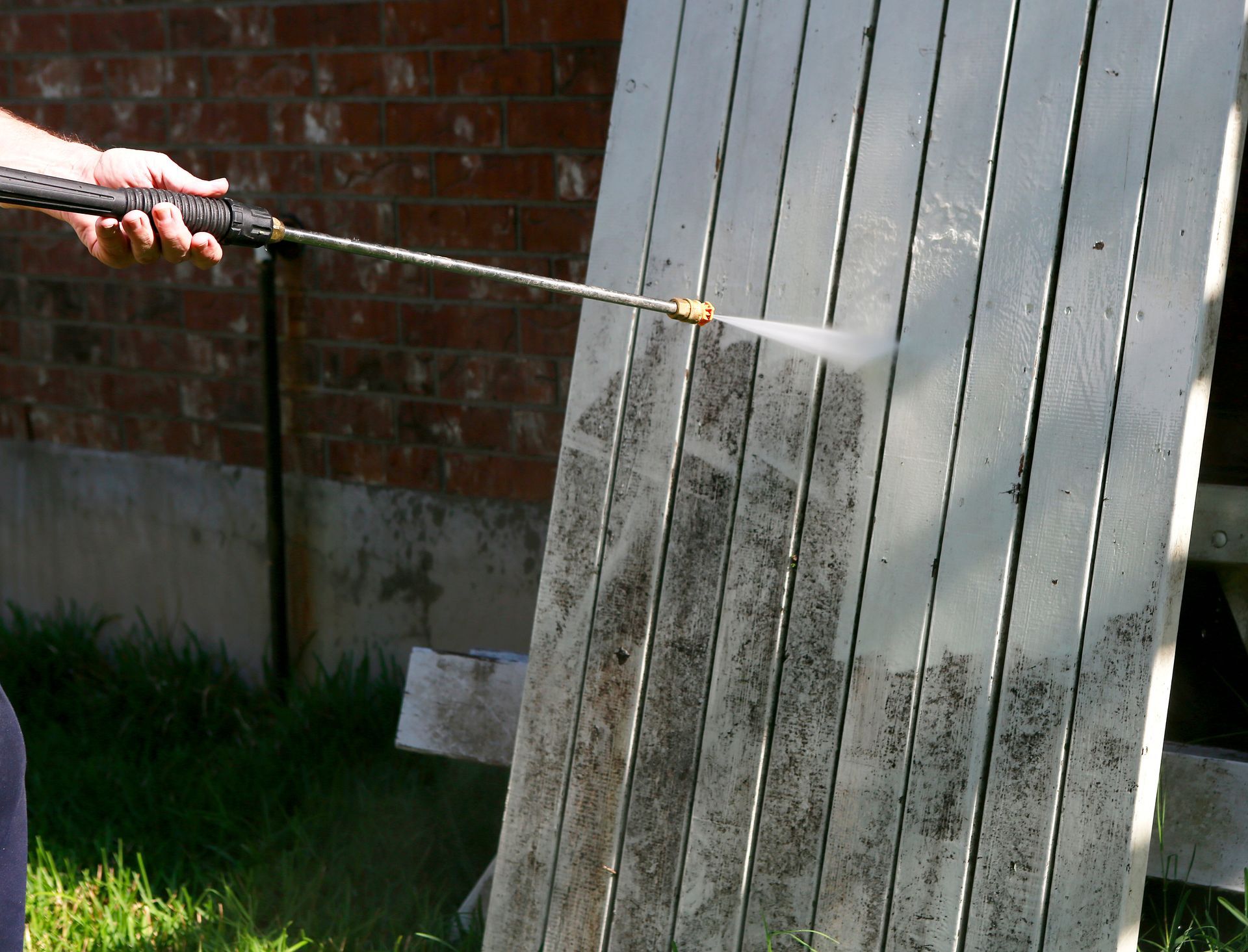 Person spraying water on a white wooden fence outdoors beside a brick wall.