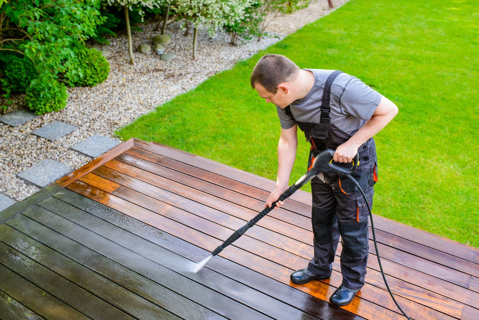 Person power-washing a wooden deck in a backyard garden.