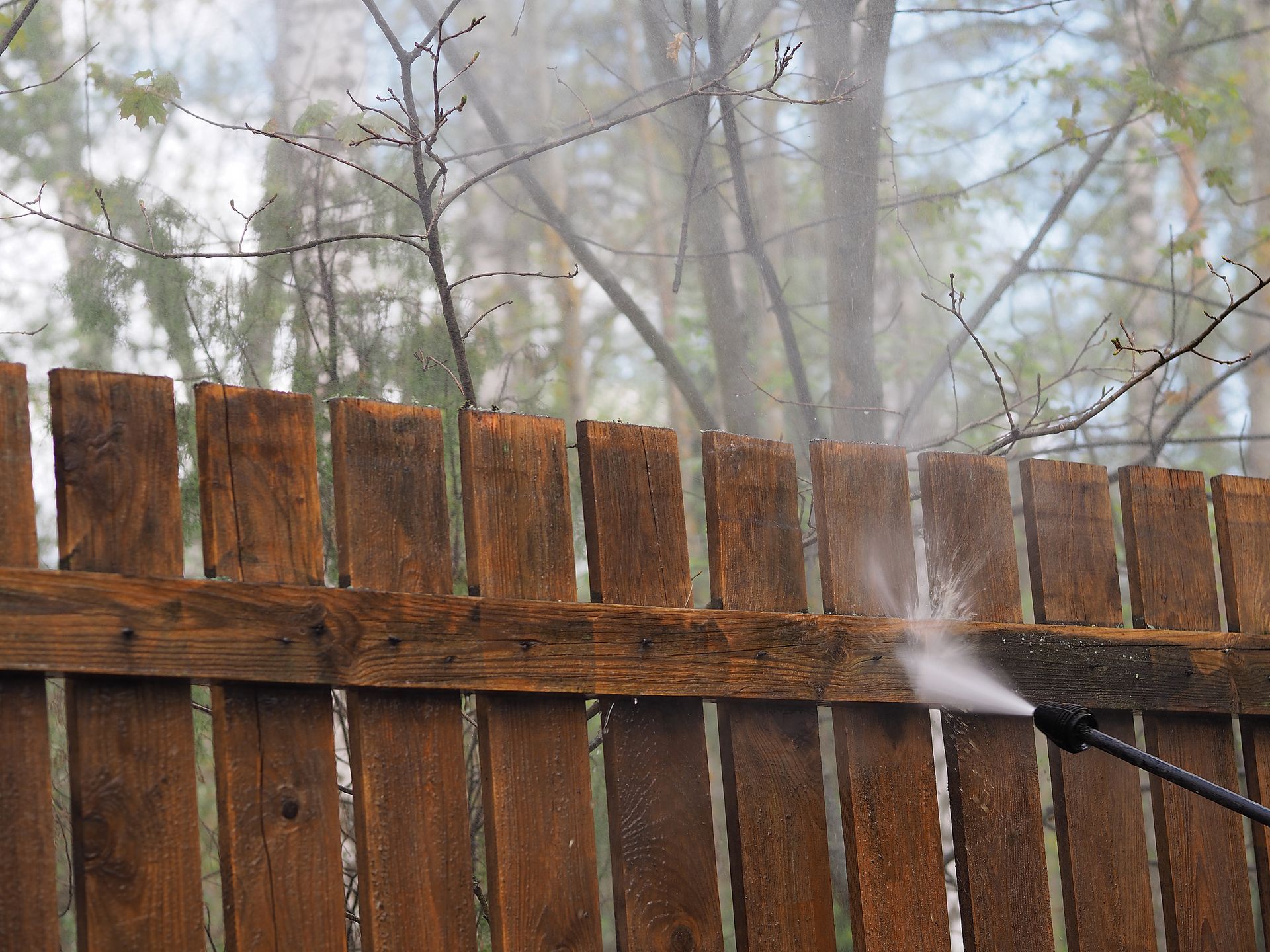 Person pressure-washes a weathered wooden fence in a misty yard.