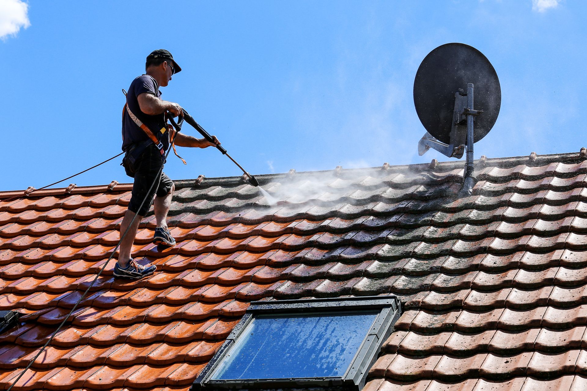 Worker pressure-washing a terracotta roof beside a satellite dish and skylight