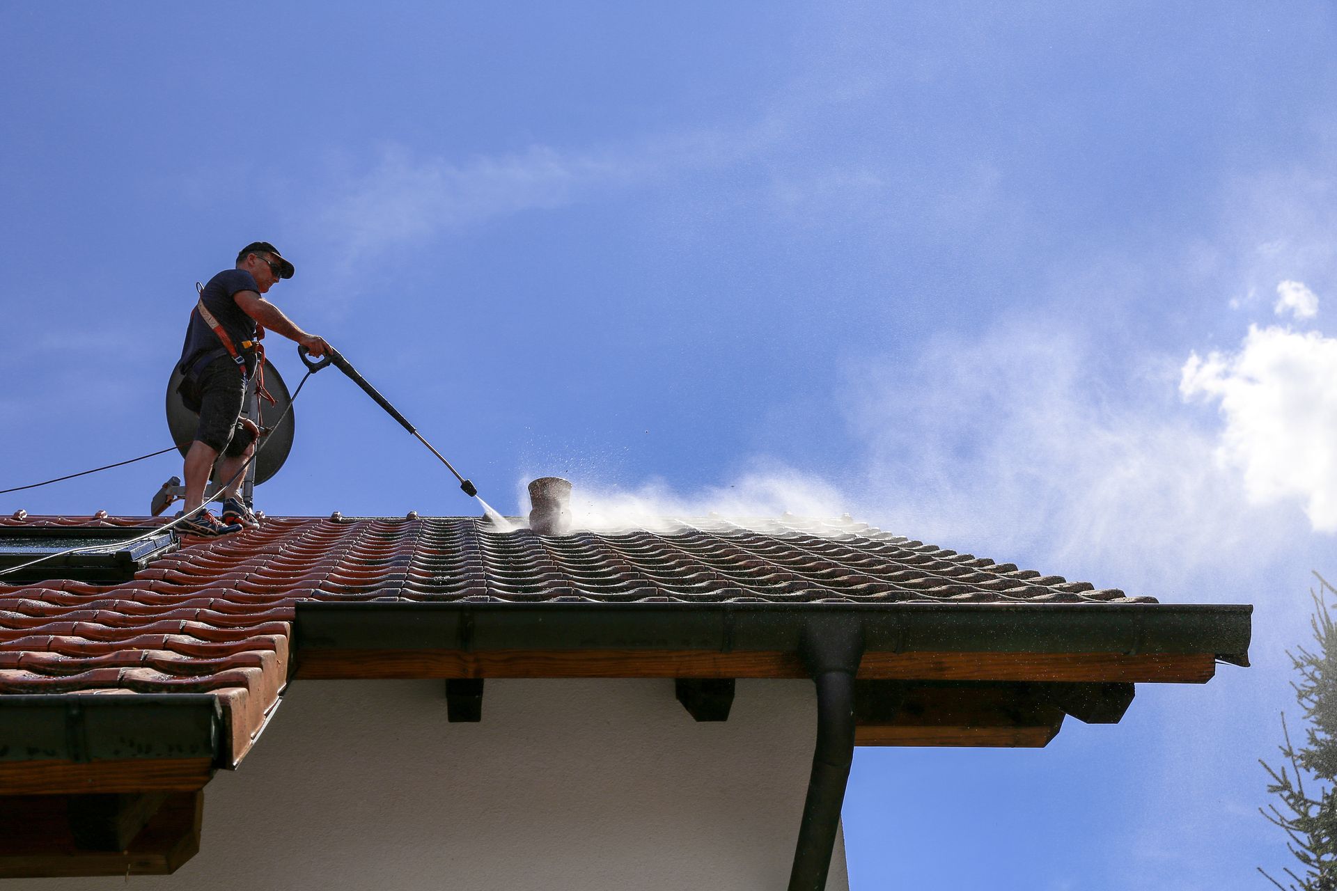 Worker pressure-washing a red-tiled roof against a blue sky