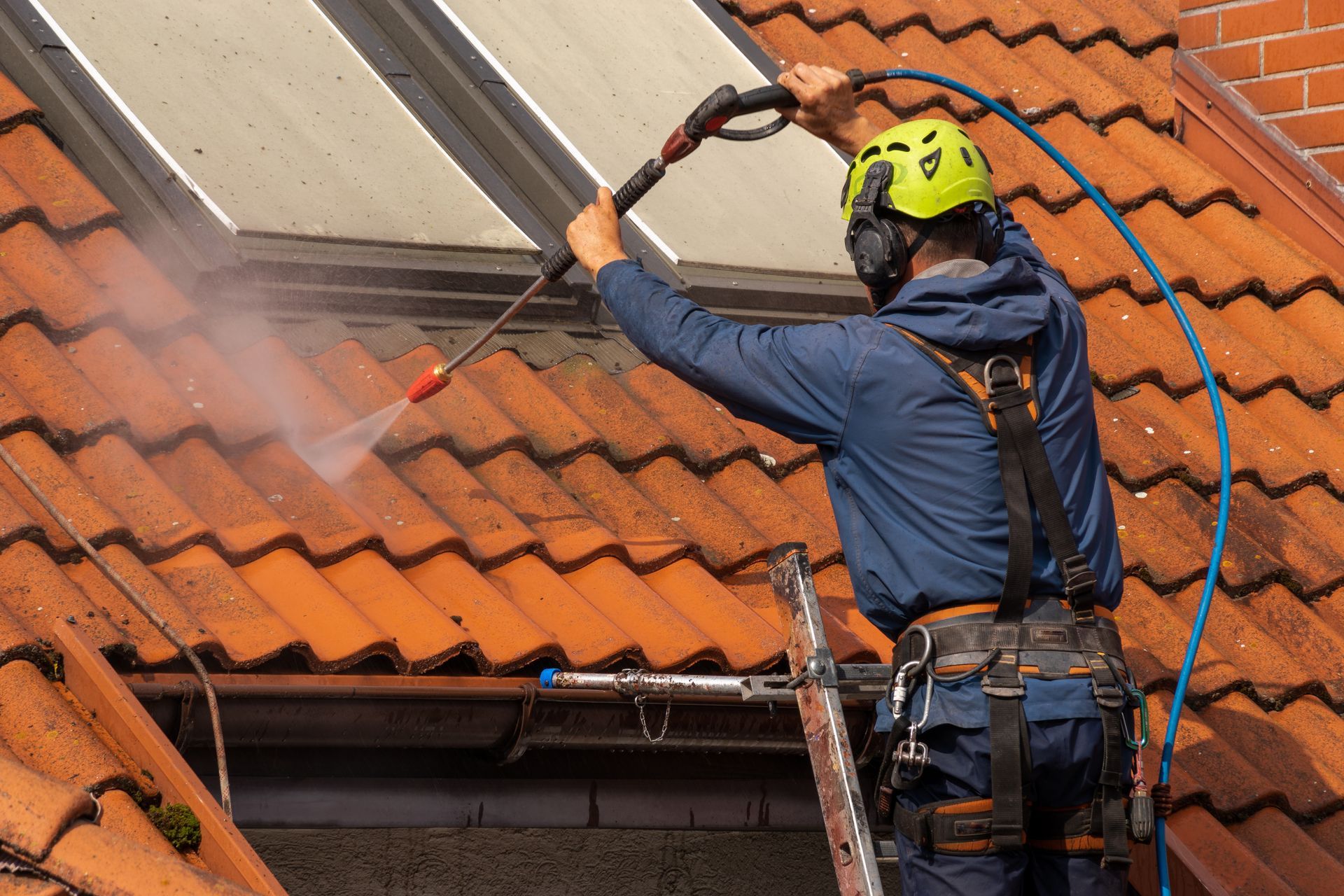 Worker pressure-washing a red tiled roof beside a skylight, wearing safety gear and harness.