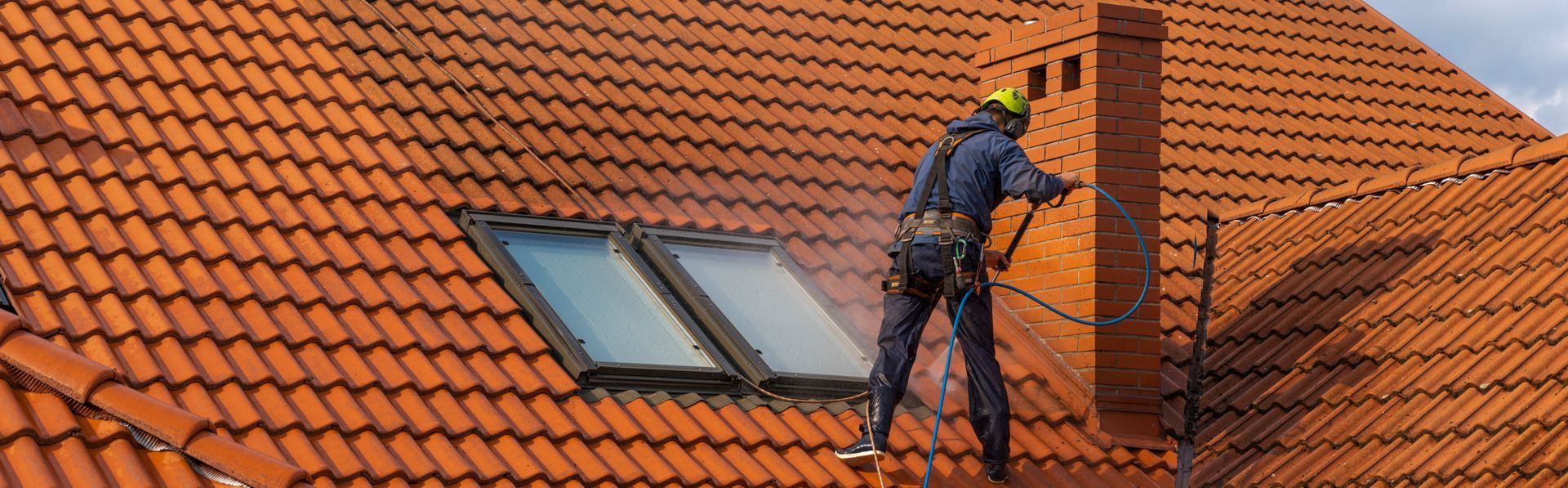Worker cleaning a red-tiled roof near a skylight, using a safety harness and hose