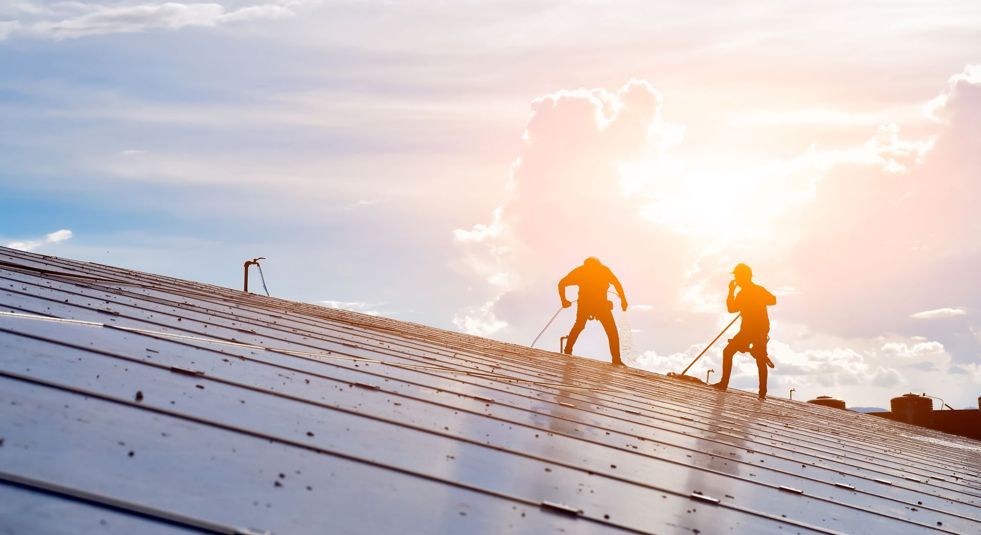 Two workers standing on a sloped metal roof at sunset, silhouetted against a bright sky.