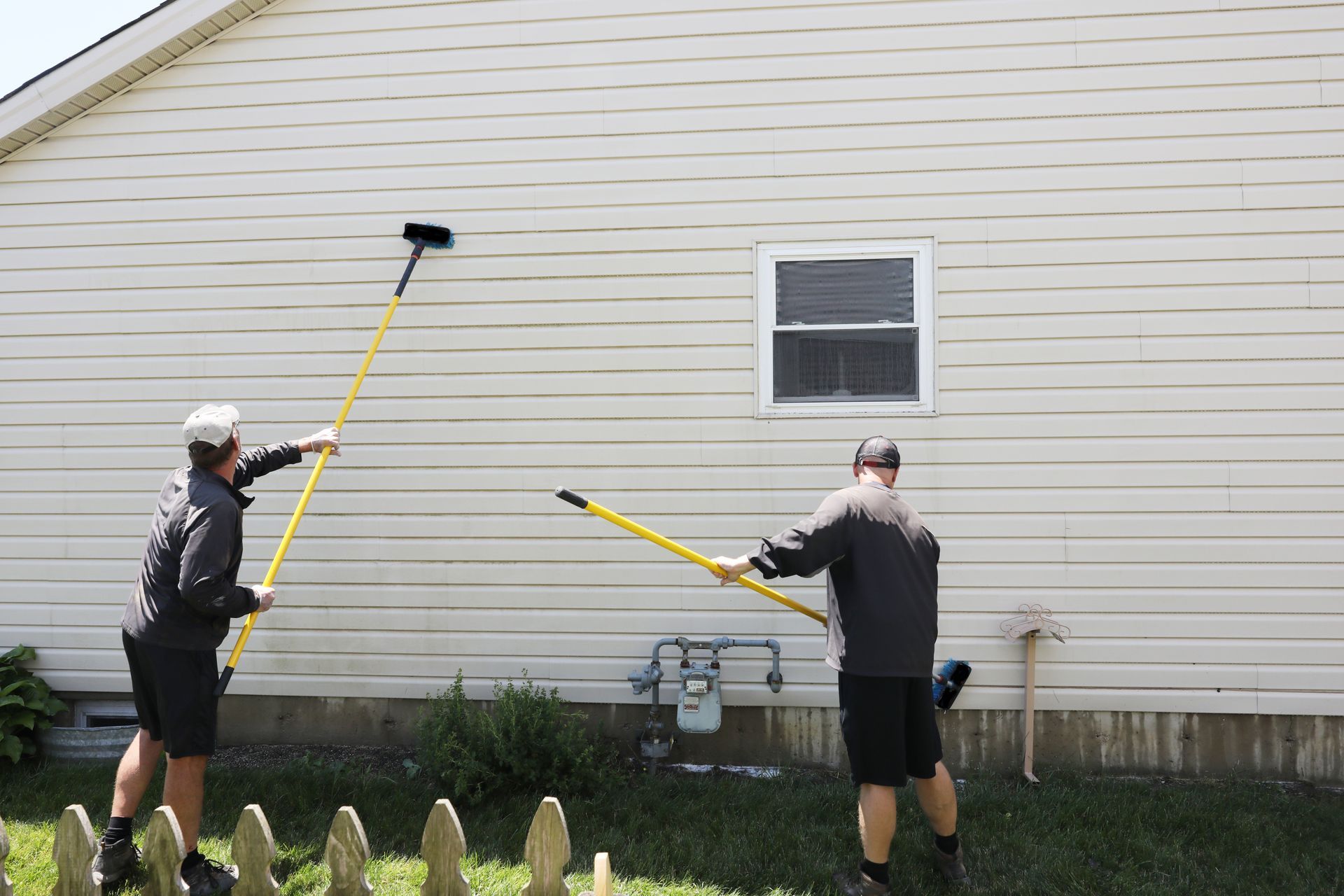 Two people using long-handled brushes to clean the side of a beige house