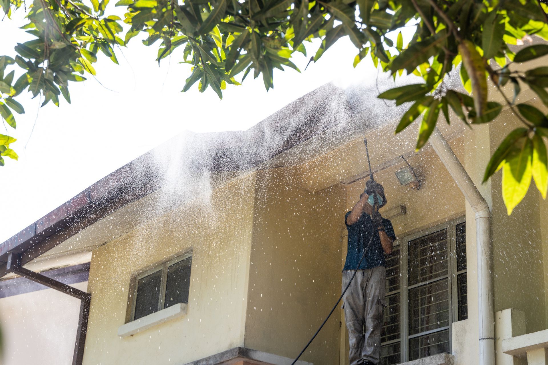 Worker pressure-washing the roof of a beige house under leafy trees