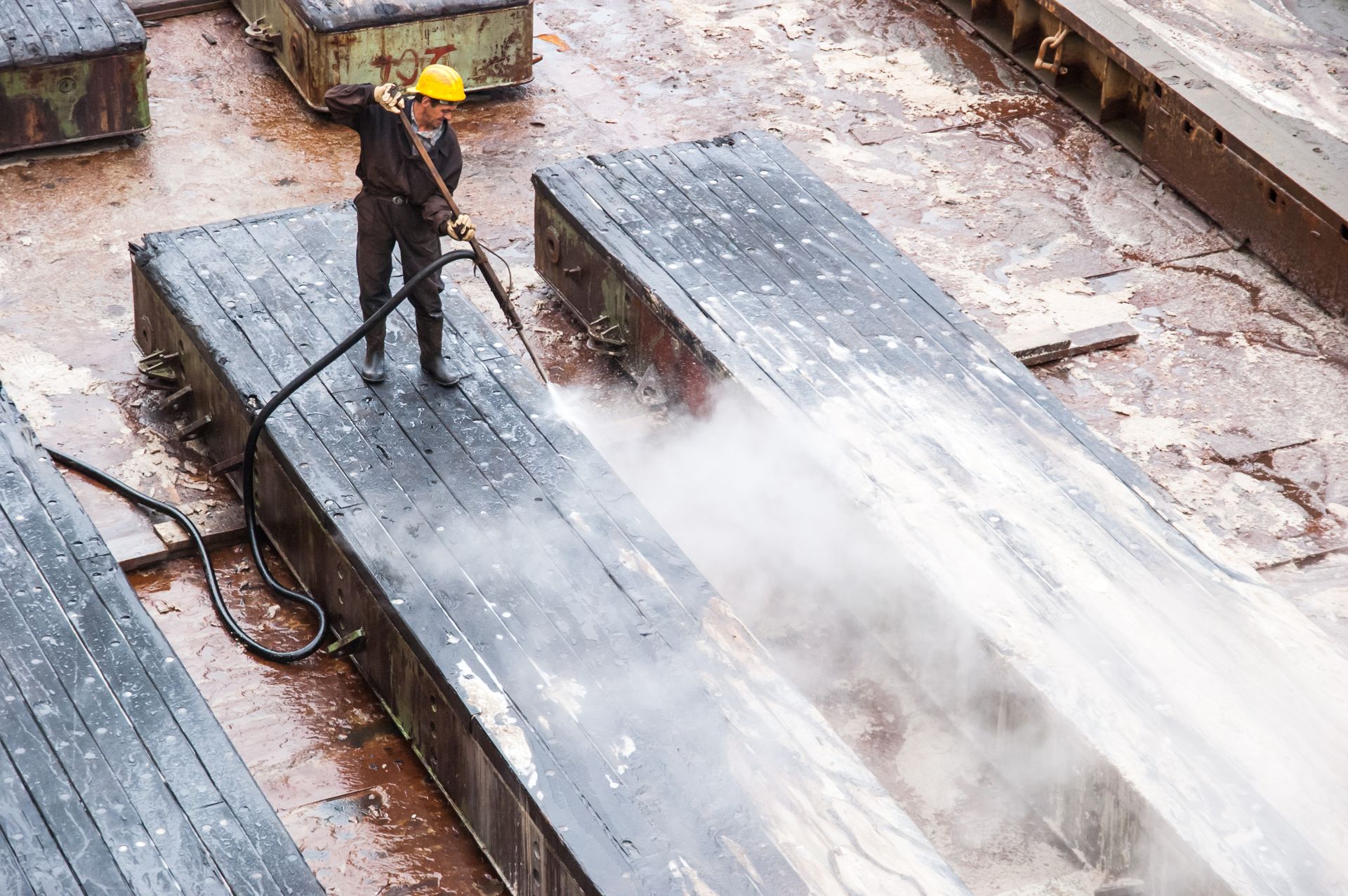 Worker in yellow hard hat pressure-washing a wet cargo deck on a ship
