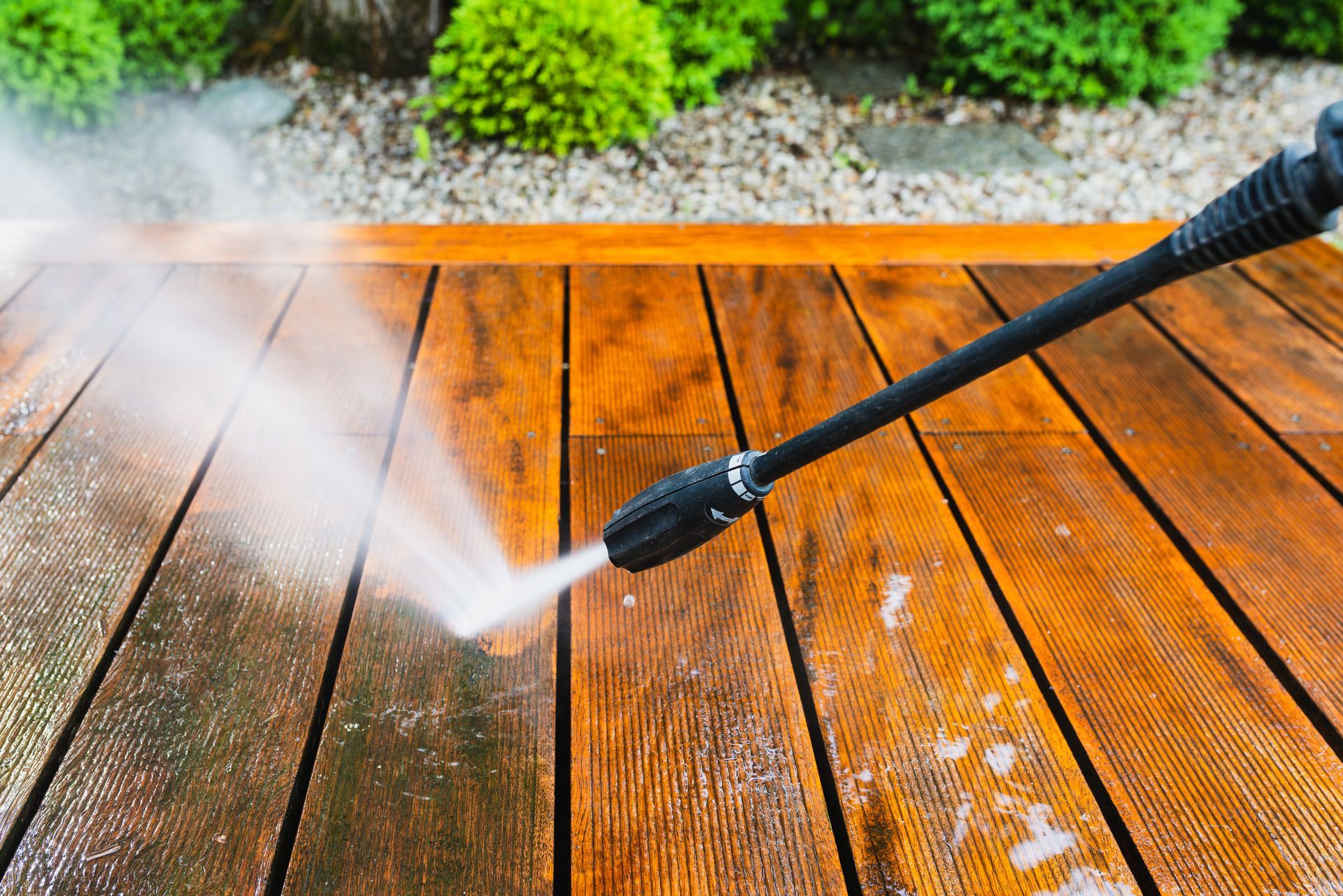 Pressure washing a wet wooden deck with green shrubs in the background