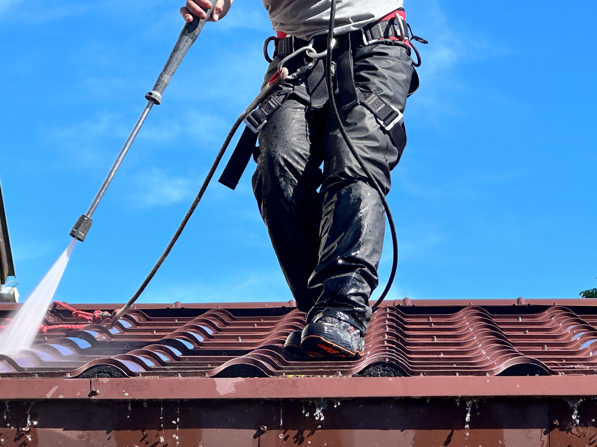 Worker pressure-washing a red tiled roof with a hose and safety harness under a blue sky