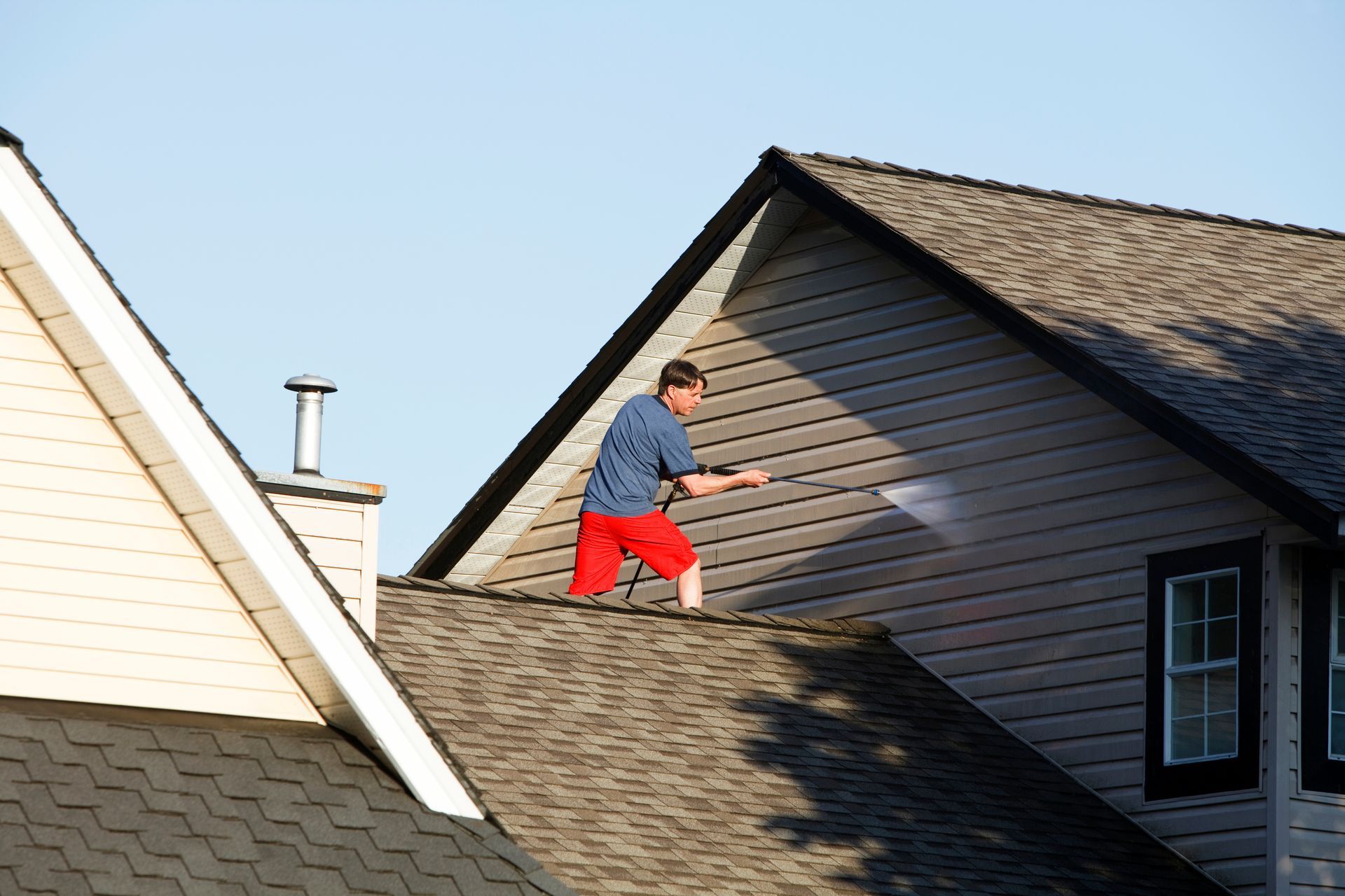 Worker in red pants on a sloped roof near a dark-sided house, using a tool in bright daylight