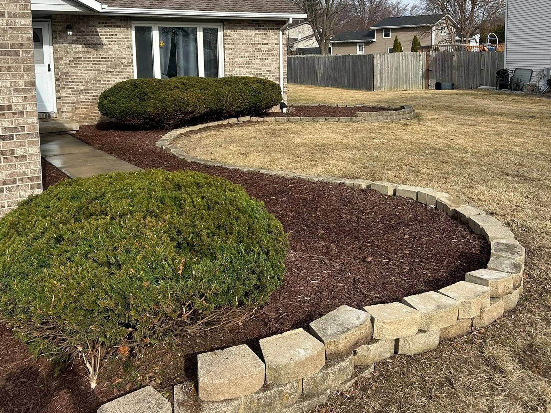 Curved stone-edged mulch bed beside a house with trimmed shrubs and a dry grassy yard