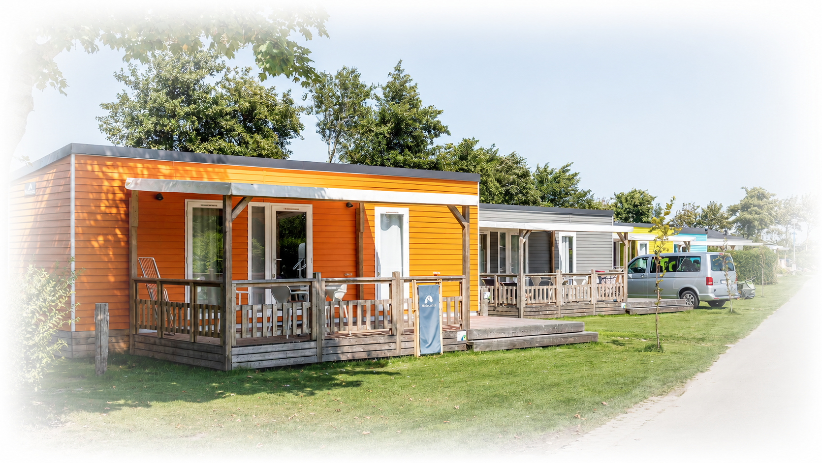 Een rij kleurrijke stacaravans en campers op een grasveld, met houten veranda's en bomen in de buurt.