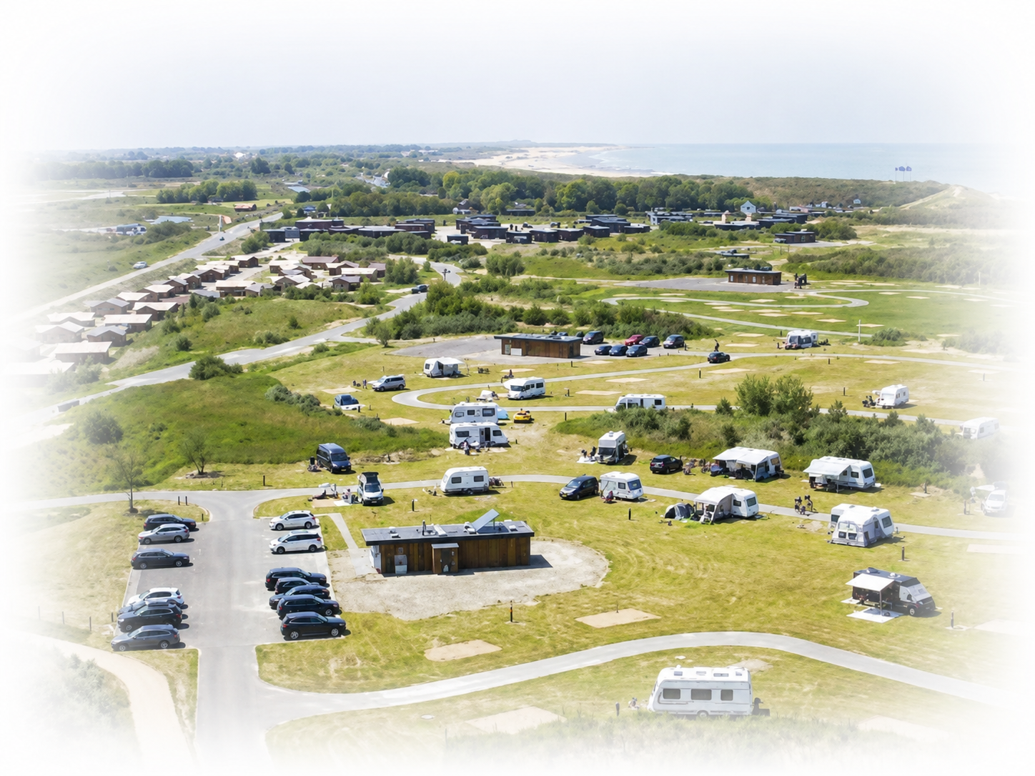 Luchtfoto van een grasrijk kampeerterrein met veel tenten, geparkeerde auto's en campers in de buurt van een weg en bomen.