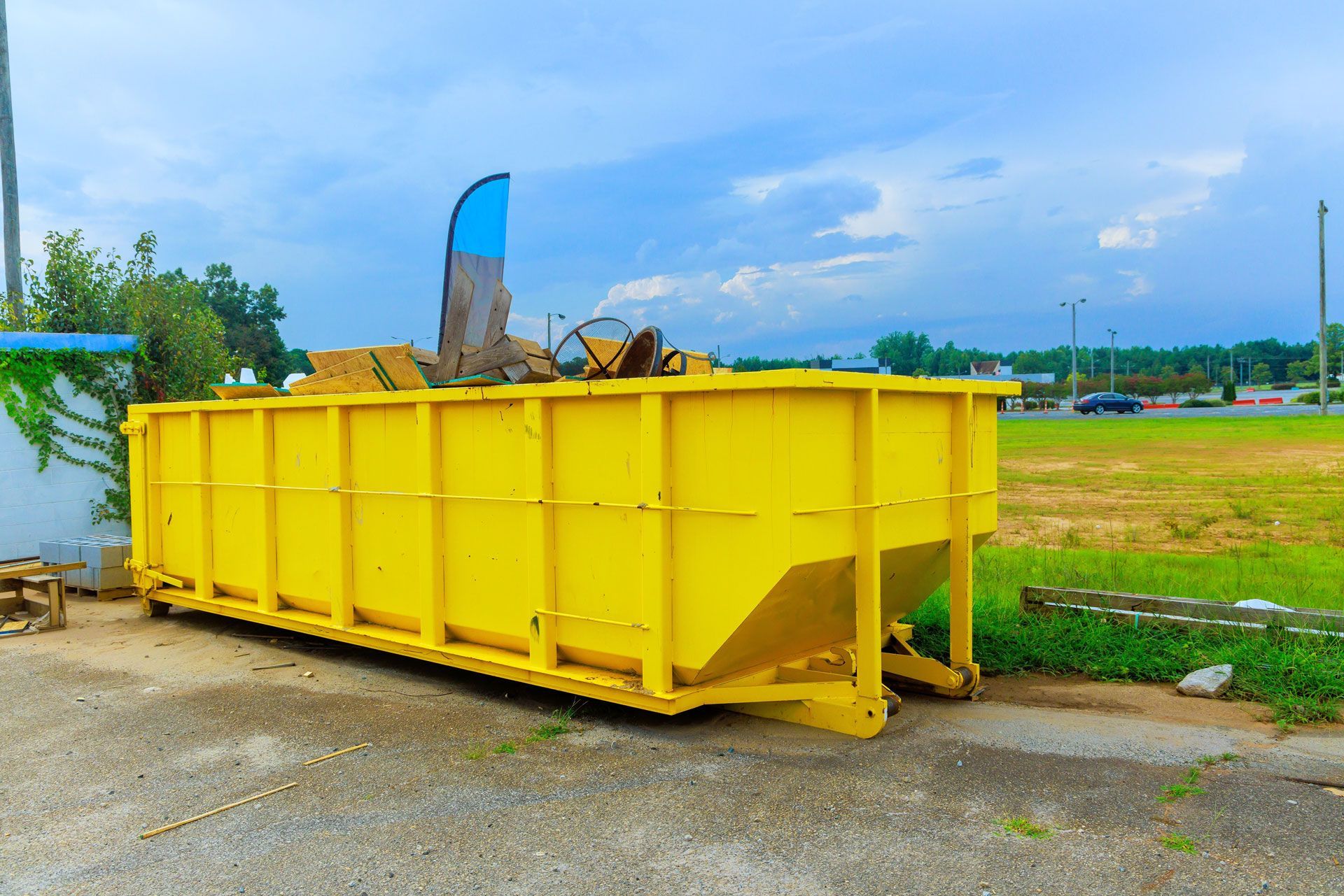Yellow dumpster, partially filled, sits outside on a gravel surface. Overcast sky in the background.
