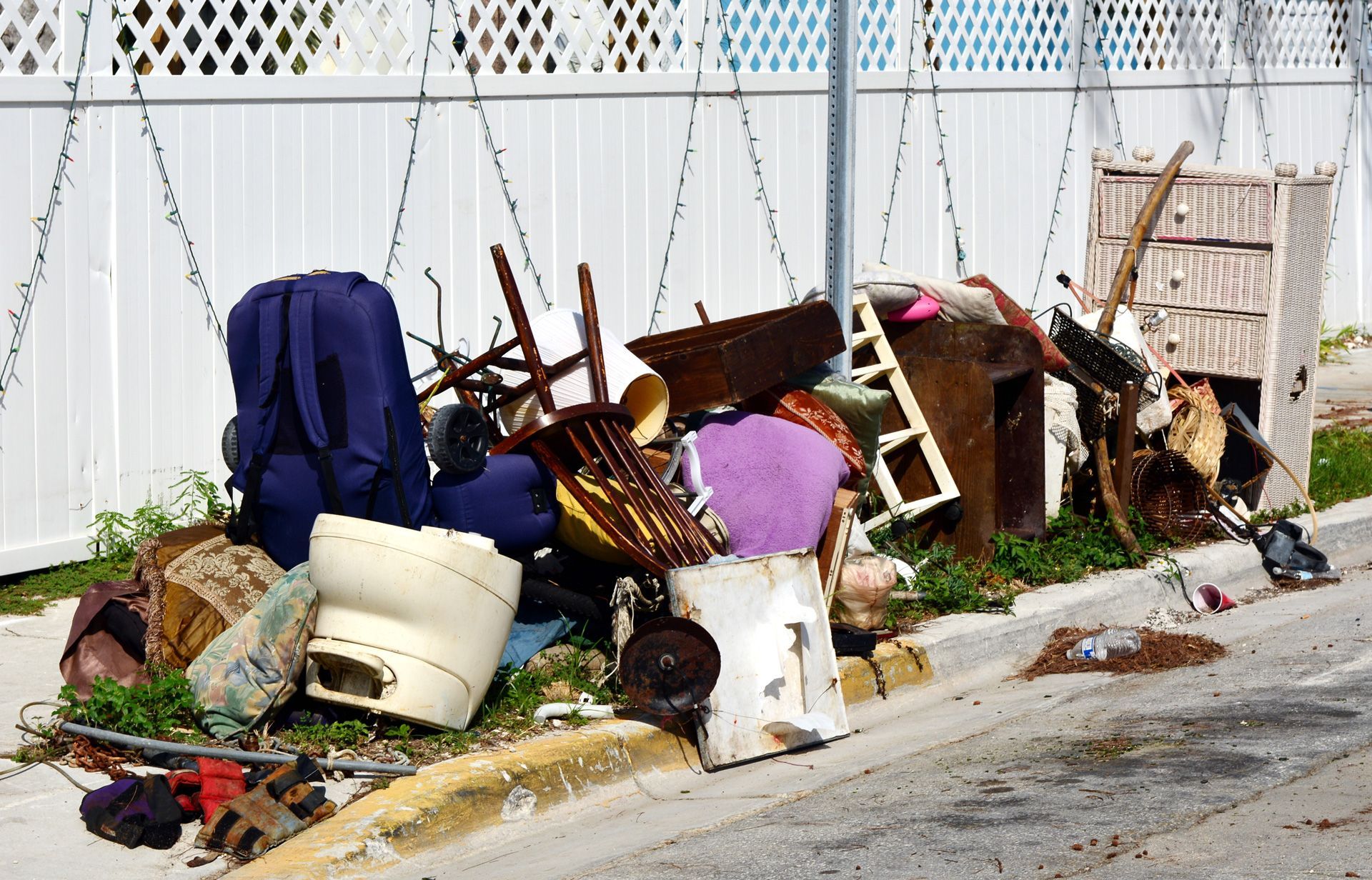 Piled curbside trash and furniture beside a white fence on a street corner