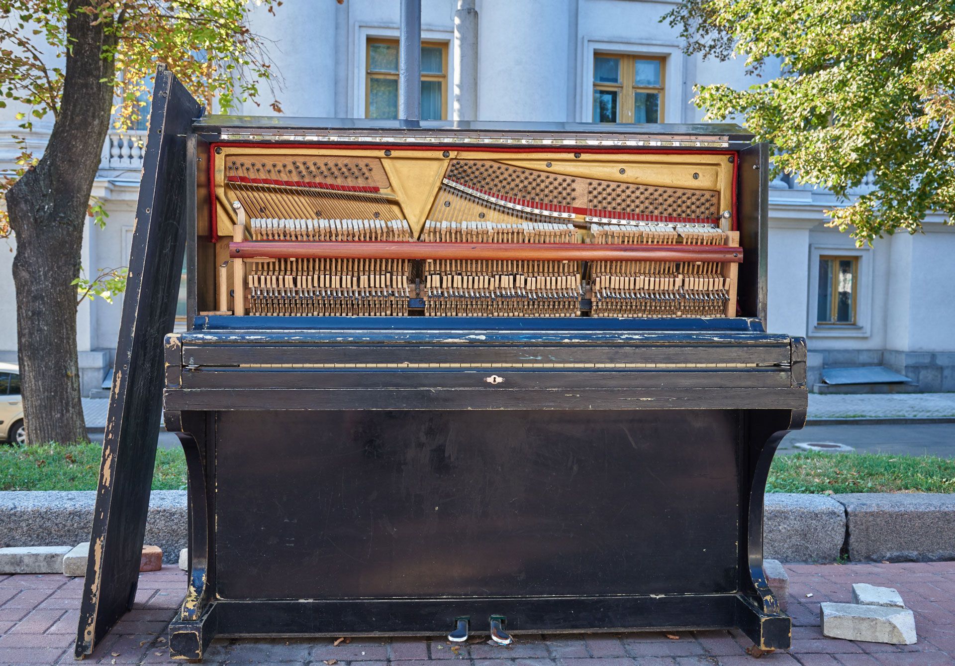 Outdoor upright piano with its top open, set on a sidewalk in front of a building.