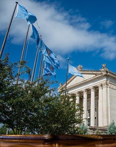 Blue flags flying beside a neoclassical building under a bright blue sky