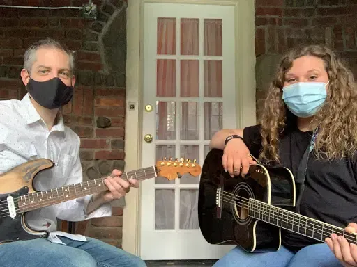 Two masked people seated with guitars on a porch, in front of a white door.